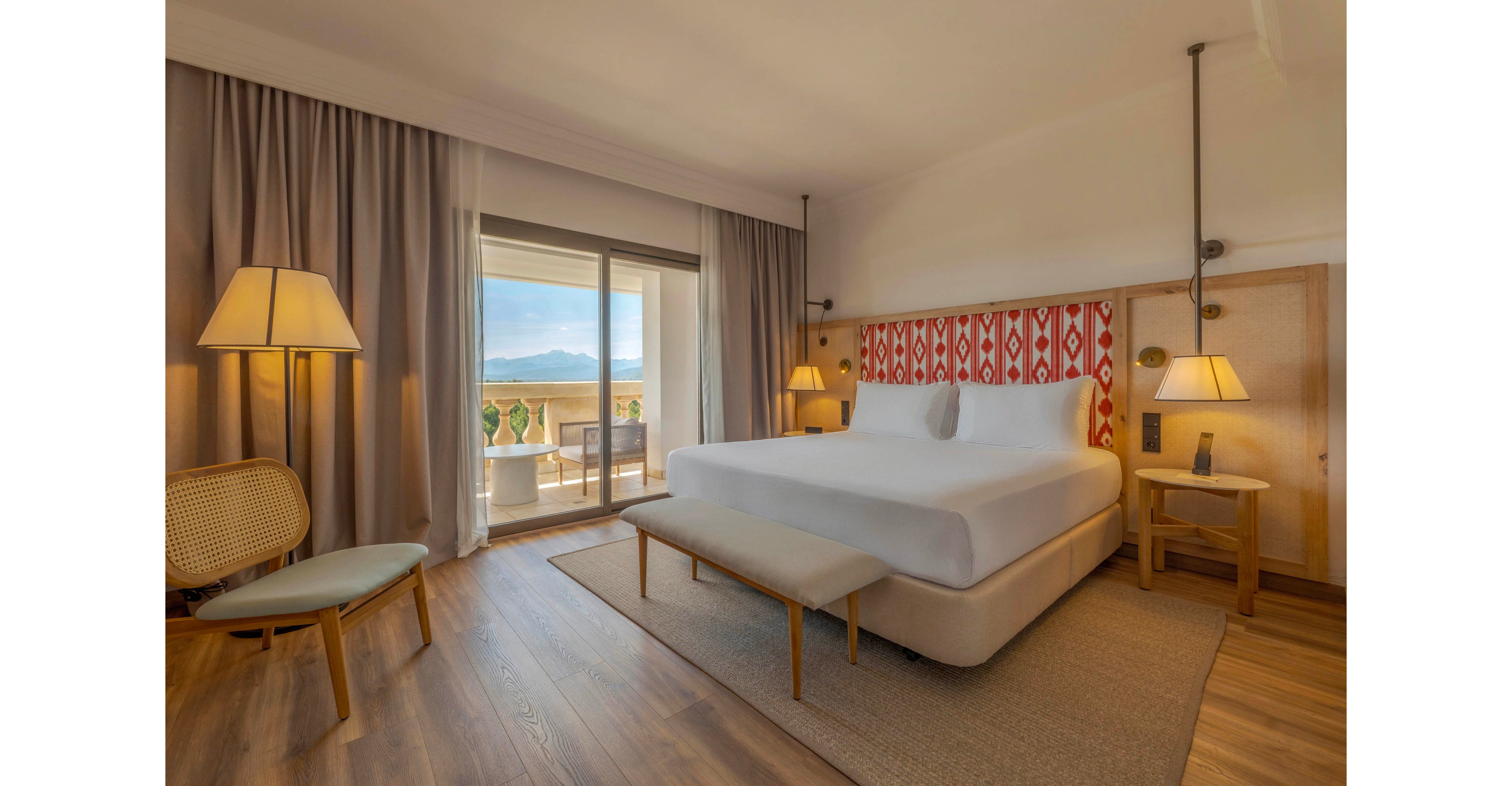 A spacious hotel bedroom features a large bed with a patterned red and white headboard and neutral bedding. Warm wooden floors, soft beige curtains, and coordinated bedside tables create a cozy, contemporary atmosphere. Multiple lamps provide ambient lighting, while a woven chair and upholstered bench add comfort and style. Sliding glass doors open to a balcony with outdoor seating and a scenic mountain view in the distance.