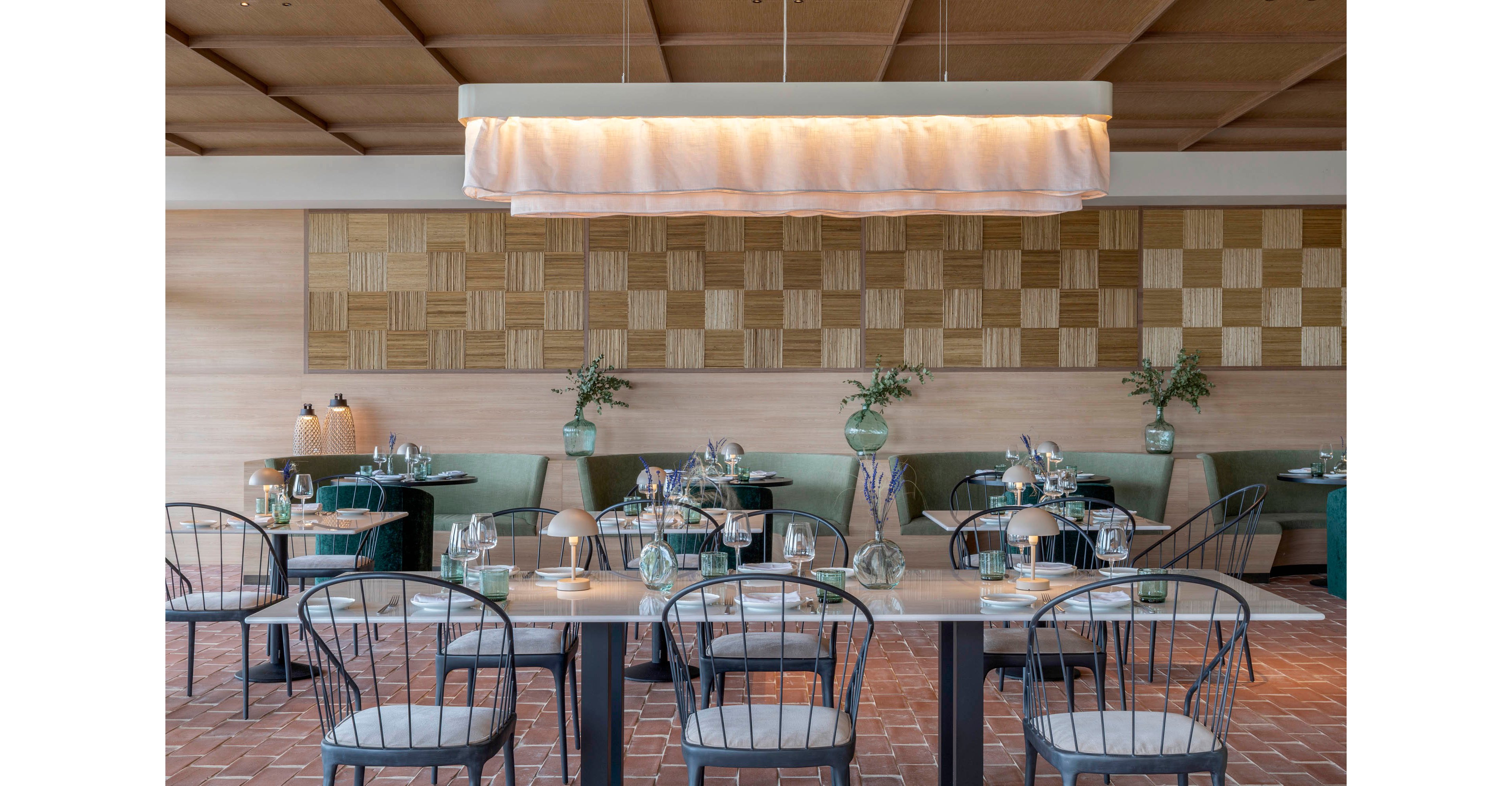 Contemporary restaurant dining room with neatly set tables and chairs. The space features a long central light fixture, patterned wood wall panels, and green upholstered banquettes. Glass vases with greenery and simple table settings create an elegant yet relaxed atmosphere. Warm lighting and natural materials emphasize a modern, minimalist design aesthetic.