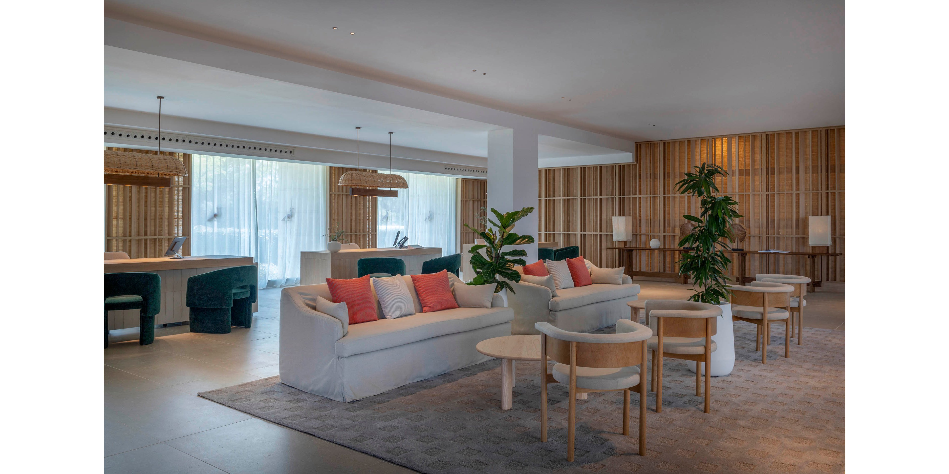 Contemporary lounge space features white sofas with coral and gray accent pillows arranged around light wood coffee tables. The setting appears to be a hotel lobby or reception area with a sleek counter and barstools in the background. Floor-to-ceiling sheer curtains diffuse natural light, while wooden slat wall panels add warmth and texture. Potted green plants and soft, neutral tones create a calm, welcoming atmosphere.