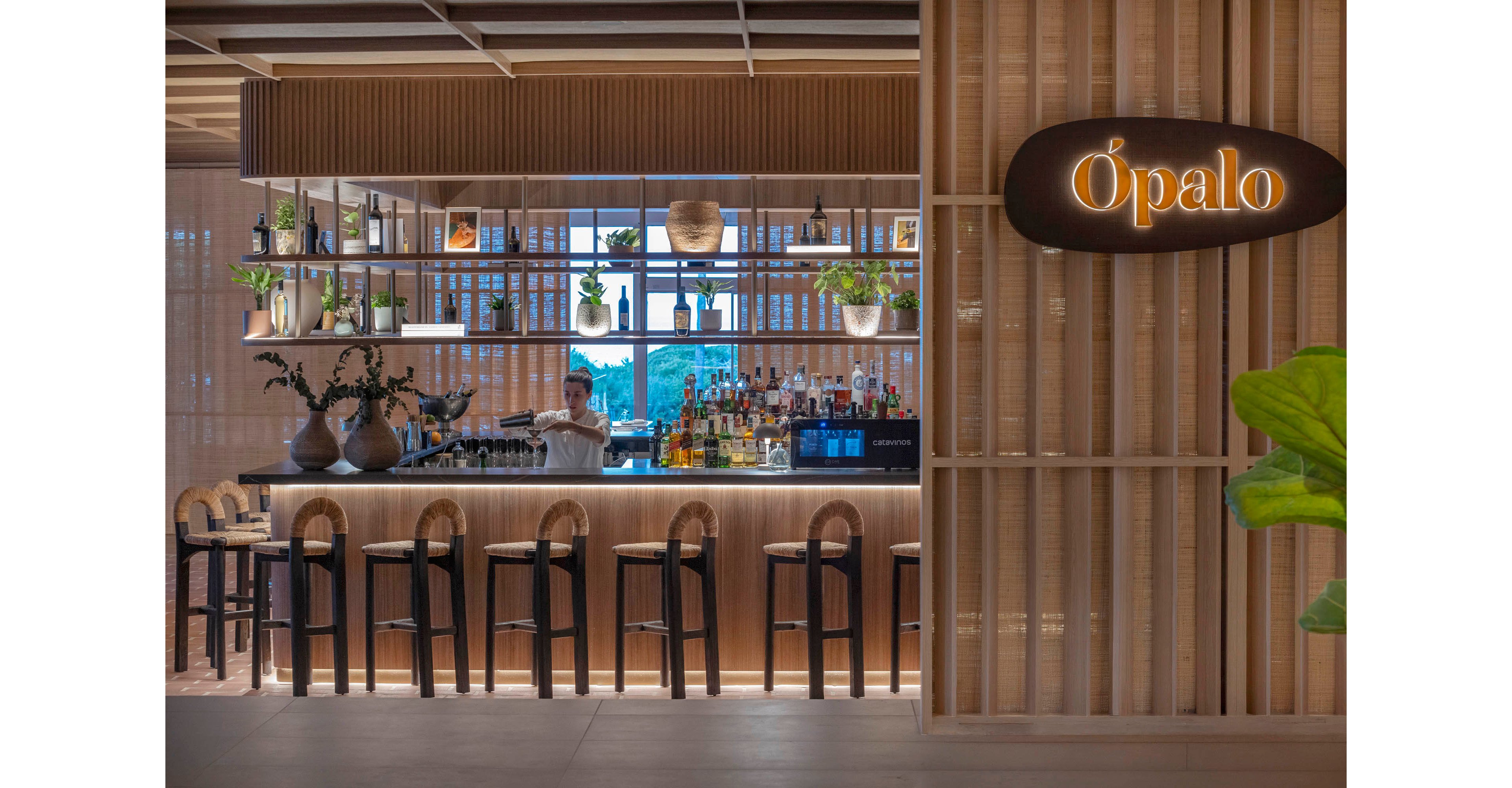 A contemporary bar named Ópalo features a sleek wooden counter with several high stools neatly arranged in front. A bartender stands behind the bar preparing drinks, surrounded by an array of bottles and glassware. Open shelving above the counter displays plants and decorative objects, adding greenery to the warm, minimalist design. Soft lighting and natural materials create a relaxed, inviting atmosphere in the stylish indoor space.