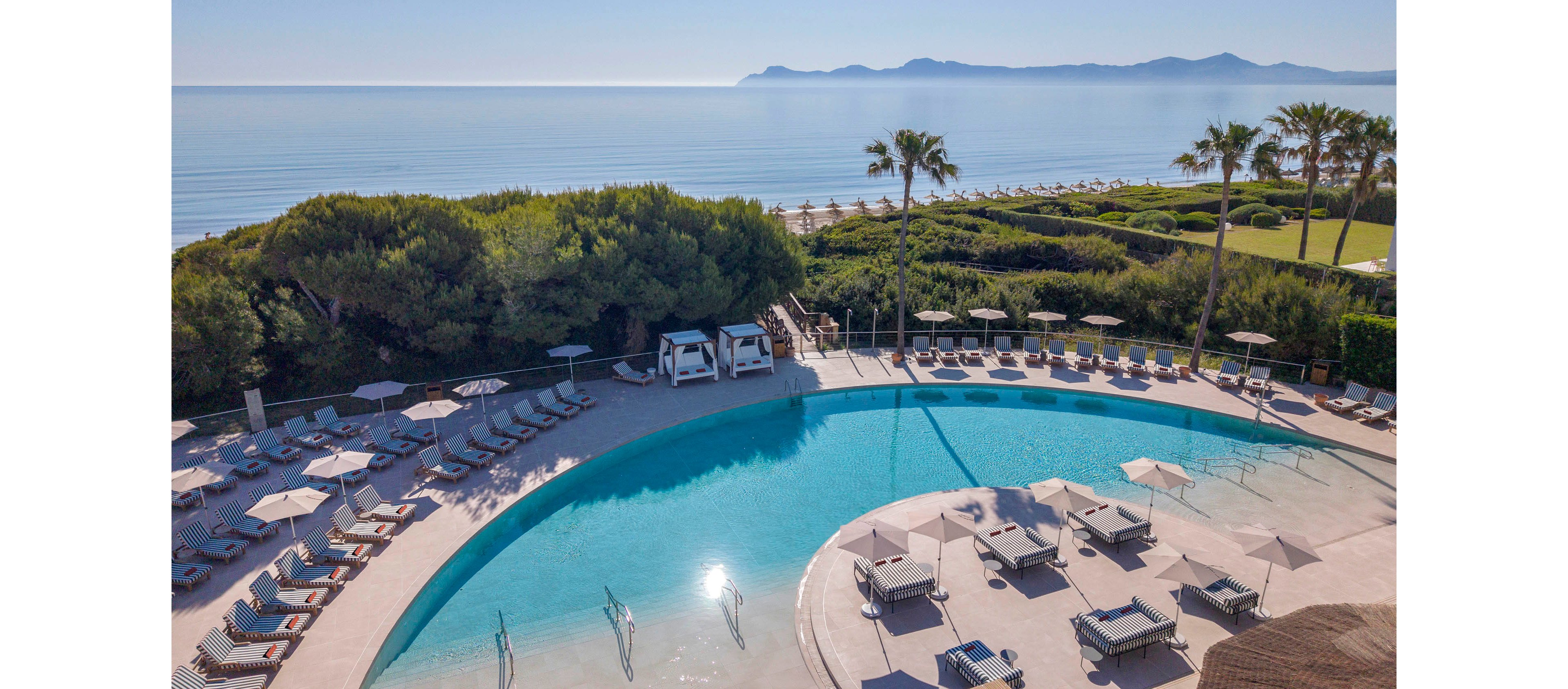 A large curved swimming pool sits at the center of a luxury beachfront resort. Sun loungers and umbrellas surround the pool, with cabanas positioned near the edge. Beyond the pool, lush greenery and palm trees frame a sandy beach leading to calm, shimmering ocean water. Distant mountains are visible on the horizon under a clear blue sky, creating a serene vacation atmosphere.
