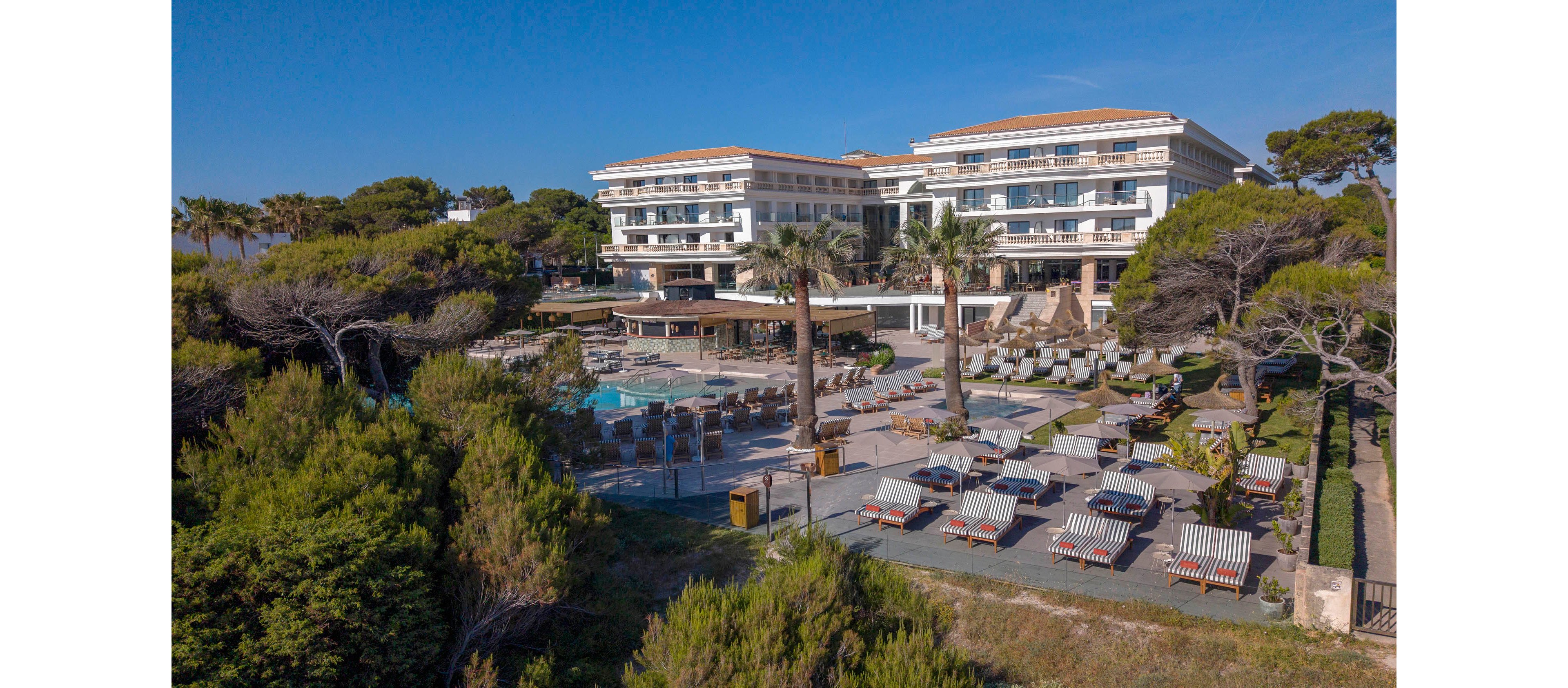 A large modern resort hotel is seen from above, surrounded by lush green trees near the coast. Multiple swimming pools, sun loungers, and umbrellas fill the outdoor terrace area in front of the building. The hotel features several stories with balconies facing the pool and garden. The overall scene suggests a tranquil vacation destination with clear weather and ample outdoor relaxation space.