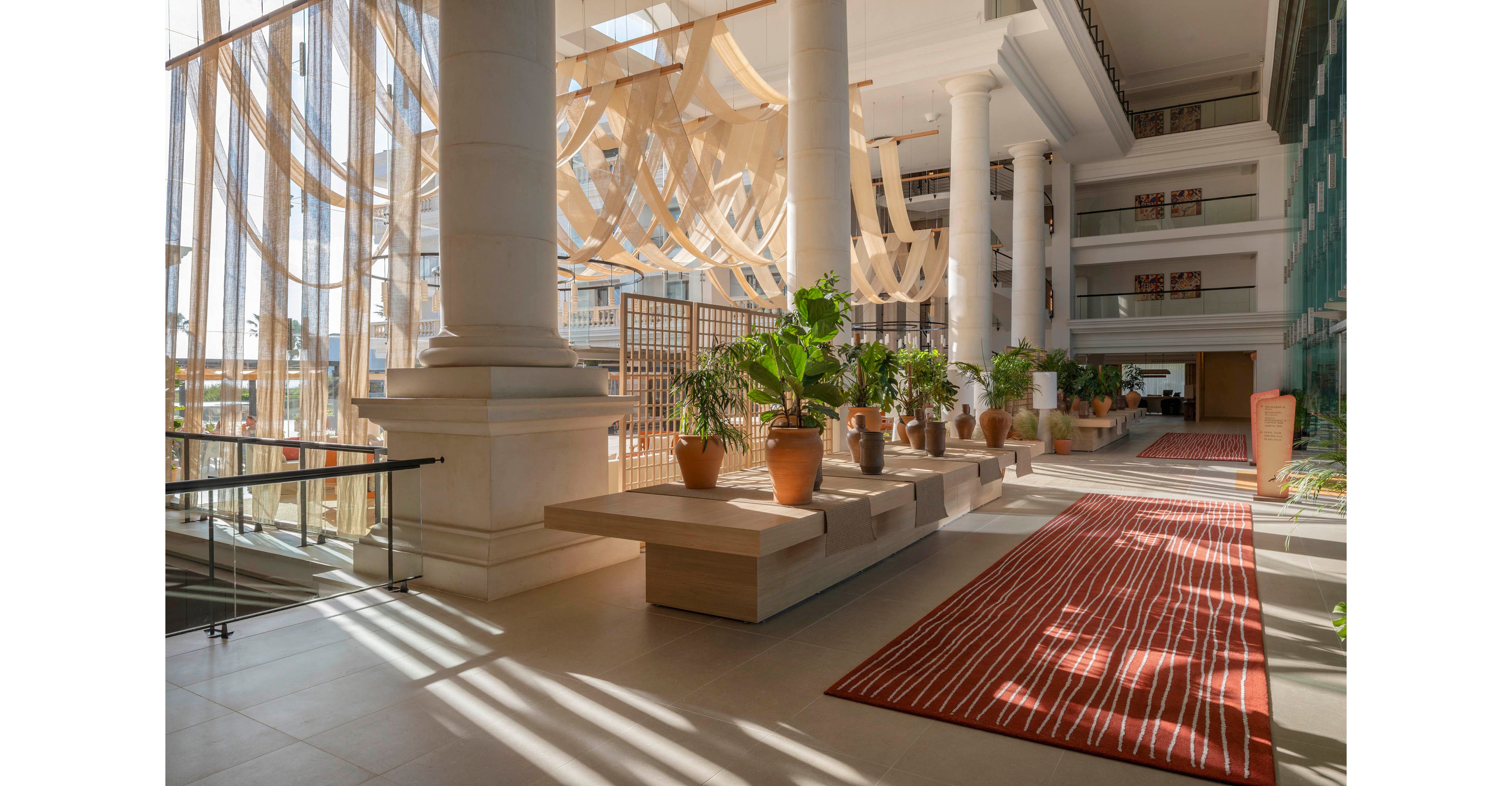 A spacious hotel atrium is filled with natural light streaming through tall windows. Large white columns frame the open area, where potted green plants sit on modern wooden platforms. A red striped runner rug leads through the space, adding contrast to the neutral tile floor. Sheer hanging fabric elements and upper-level balconies contribute to the airy, contemporary interior design.