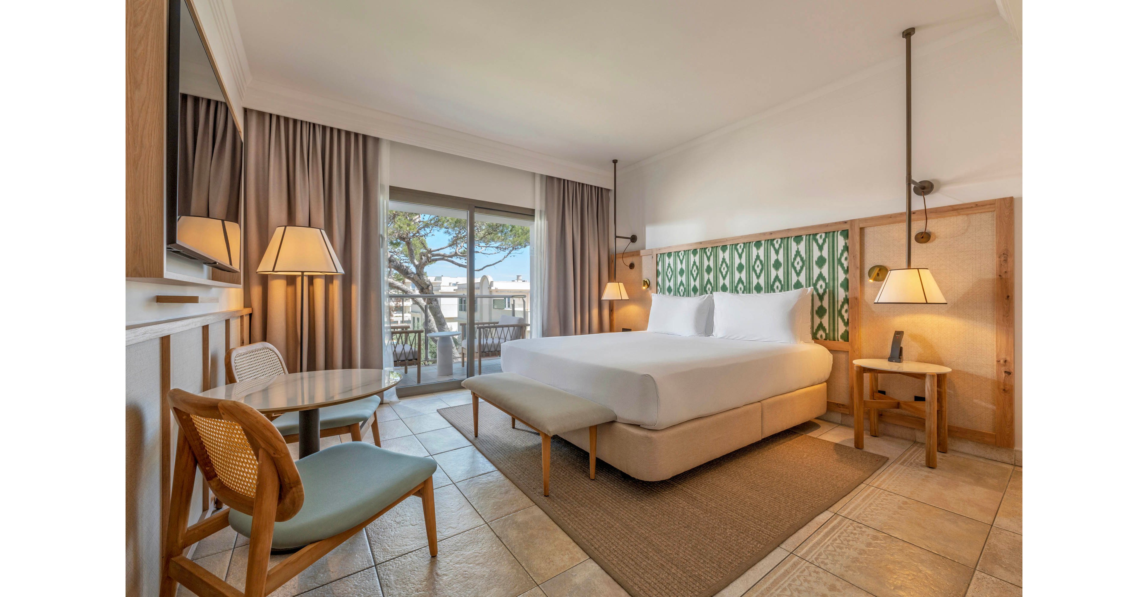 Spacious contemporary hotel room featuring a large bed with white linens and a cushioned bench at the foot. The room includes a small round table with chairs, bedside tables, and warm pendant lighting. Neutral tones and patterned green headboard accents create a calm, inviting atmosphere. Large sliding glass doors open to a balcony with outdoor seating and a view of trees and nearby buildings.