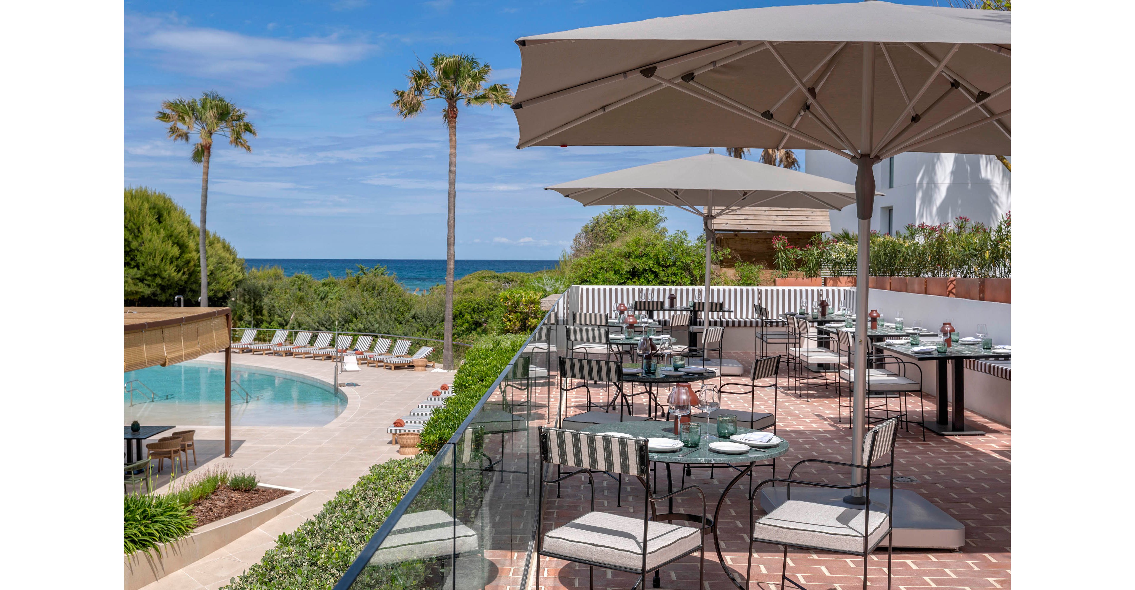 An outdoor hotel terrace dining area overlooks a swimming pool and the sea. Tables are neatly set with plates and glasses under large sun umbrellas, creating a relaxed al fresco atmosphere. Lounge chairs line the curved pool beside landscaped greenery and palm trees. The bright daylight and clear sky emphasize a calm, upscale coastal resort setting.