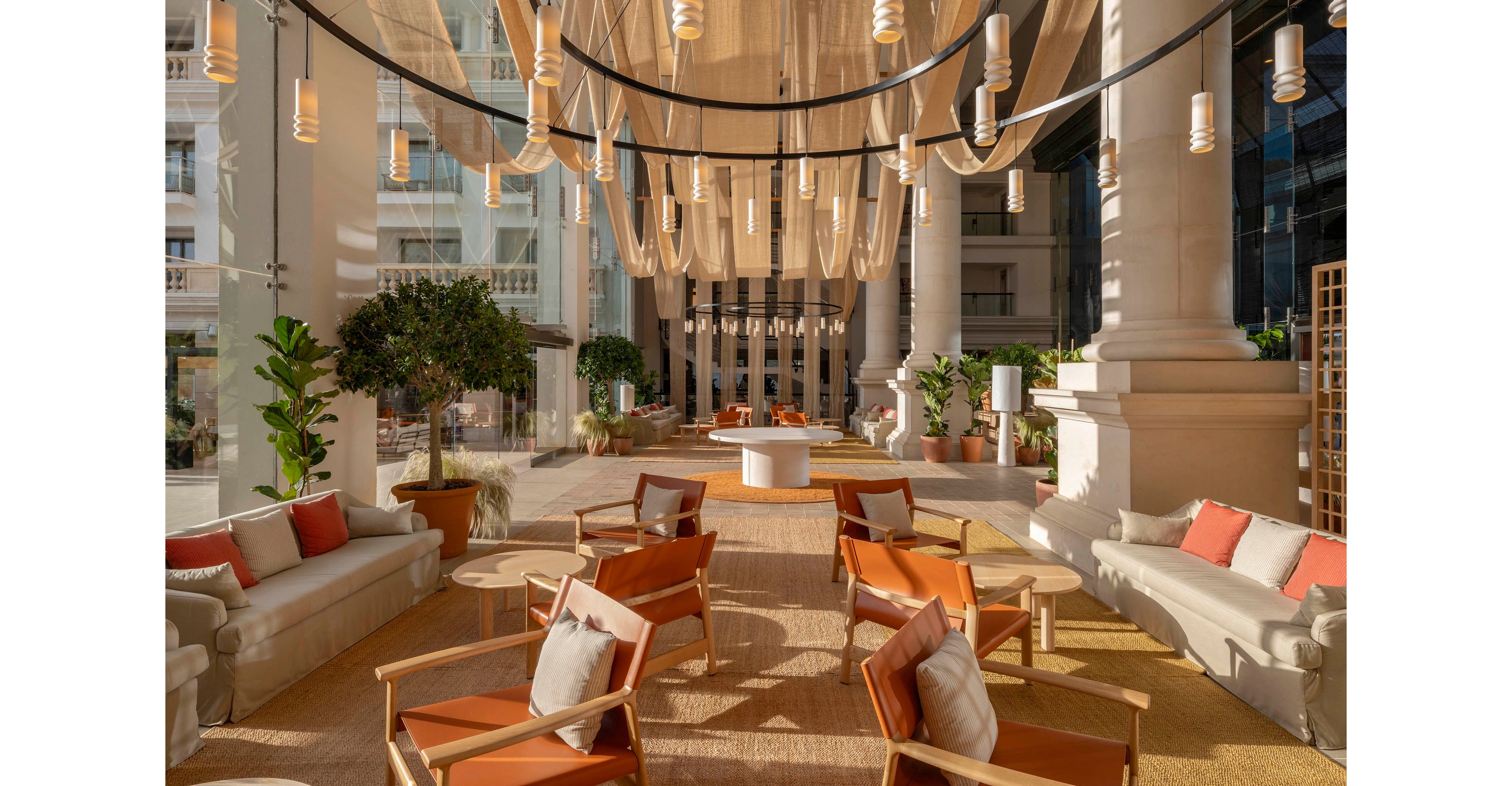 A spacious hotel lobby lounge features modern armchairs and sofas arranged in conversational groupings. Warm sunlight streams through large glass windows, illuminating neutral textiles and terracotta-colored cushions. Sheer fabric panels and hanging pendant lights create a sculptural ceiling focal point. Potted plants and natural wood tables add a relaxed, resort-like atmosphere to the elegant interior.