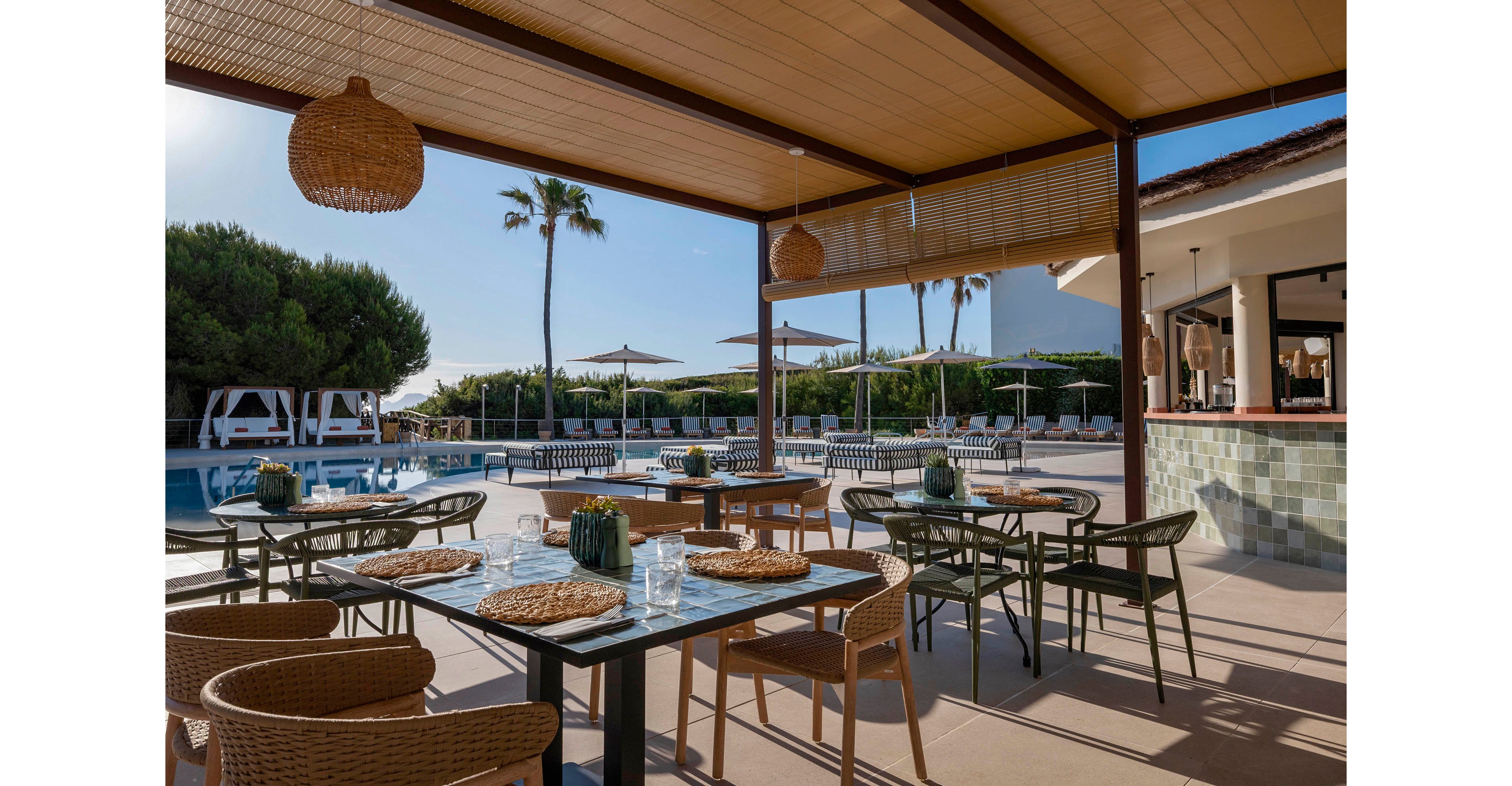 An open-air restaurant terrace overlooks a serene swimming pool on a sunny day. Wicker chairs and glass-topped tables with woven placemats are arranged neatly under a wooden pergola. In the background, sun loungers, umbrellas, and cabanas line the poolside area, framed by lush greenery and tall palm trees. Warm natural light and neutral tones create a relaxed, resort-style atmosphere ideal for vacation dining.