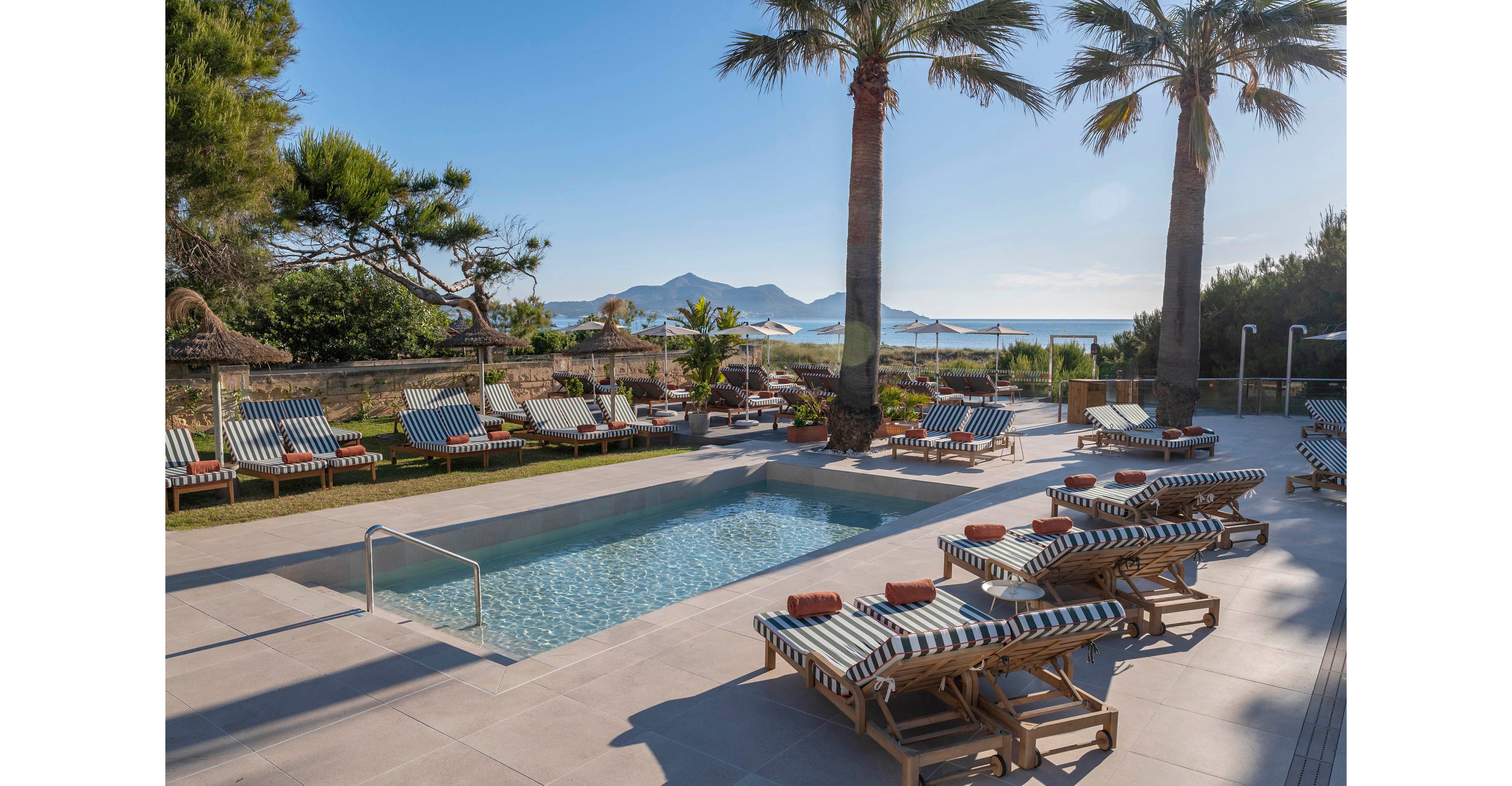 An outdoor swimming pool is surrounded by striped sun loungers and palm trees at a coastal resort. Rolled towels rest on the loungers, ready for guests to relax by the water. In the background, parasols and additional seating overlook the sea and distant mountains. The scene is bright and tranquil, suggesting a warm, sunny vacation atmosphere.