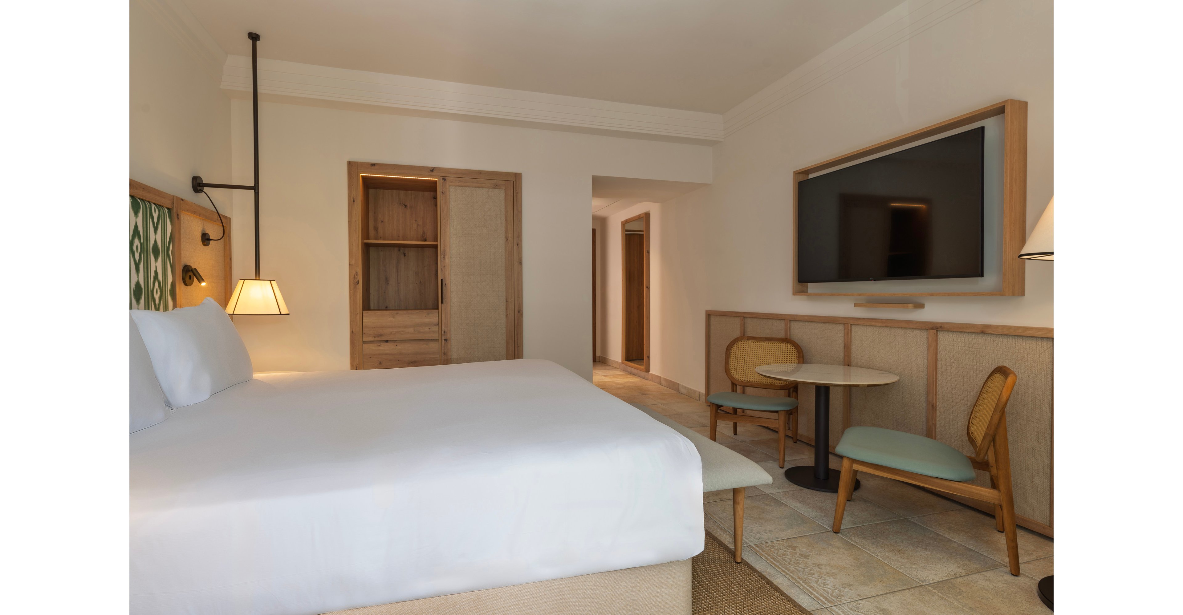 A contemporary hotel room features a large neatly made bed with white linens in the foreground. On the right side, a flat-screen television is mounted above a light wood panel. A small round table with two chairs sits beside the wall, creating a cozy seating area. In the background, wooden sliding doors reveal a closet space, and a hallway leads toward the entrance, all in neutral tones for a calm atmosphere.