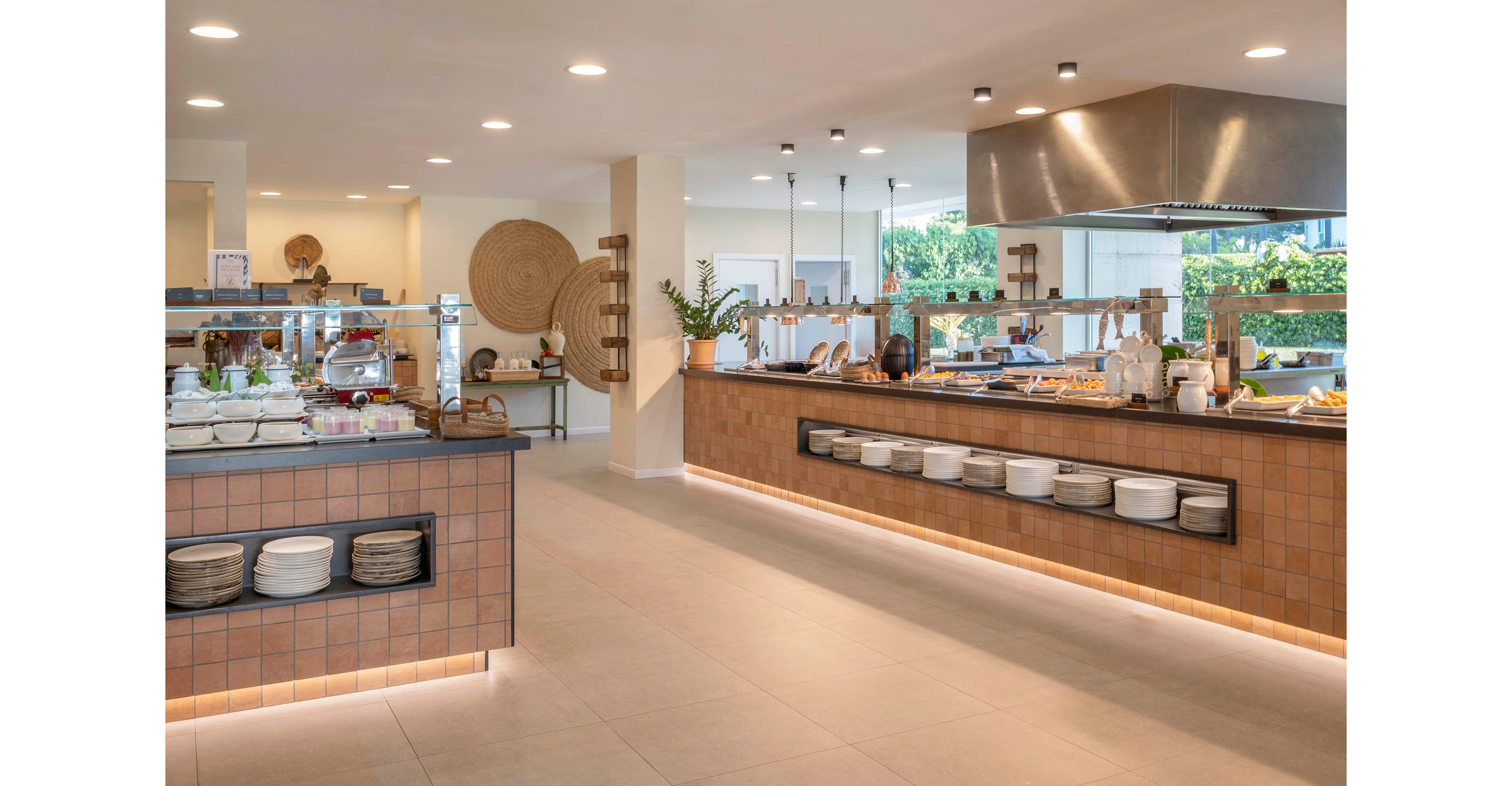 Spacious self-service buffet area in a modern restaurant with tiled counters and open shelving. Stainless steel heat lamps and a large ventilation hood hang above the main food station. Plates, bowls, and serving dishes are neatly arranged along the counters, with decorative plants and woven wall art adding warmth. Large windows in the background bring in natural light and views of greenery, creating an inviting dining atmosphere.