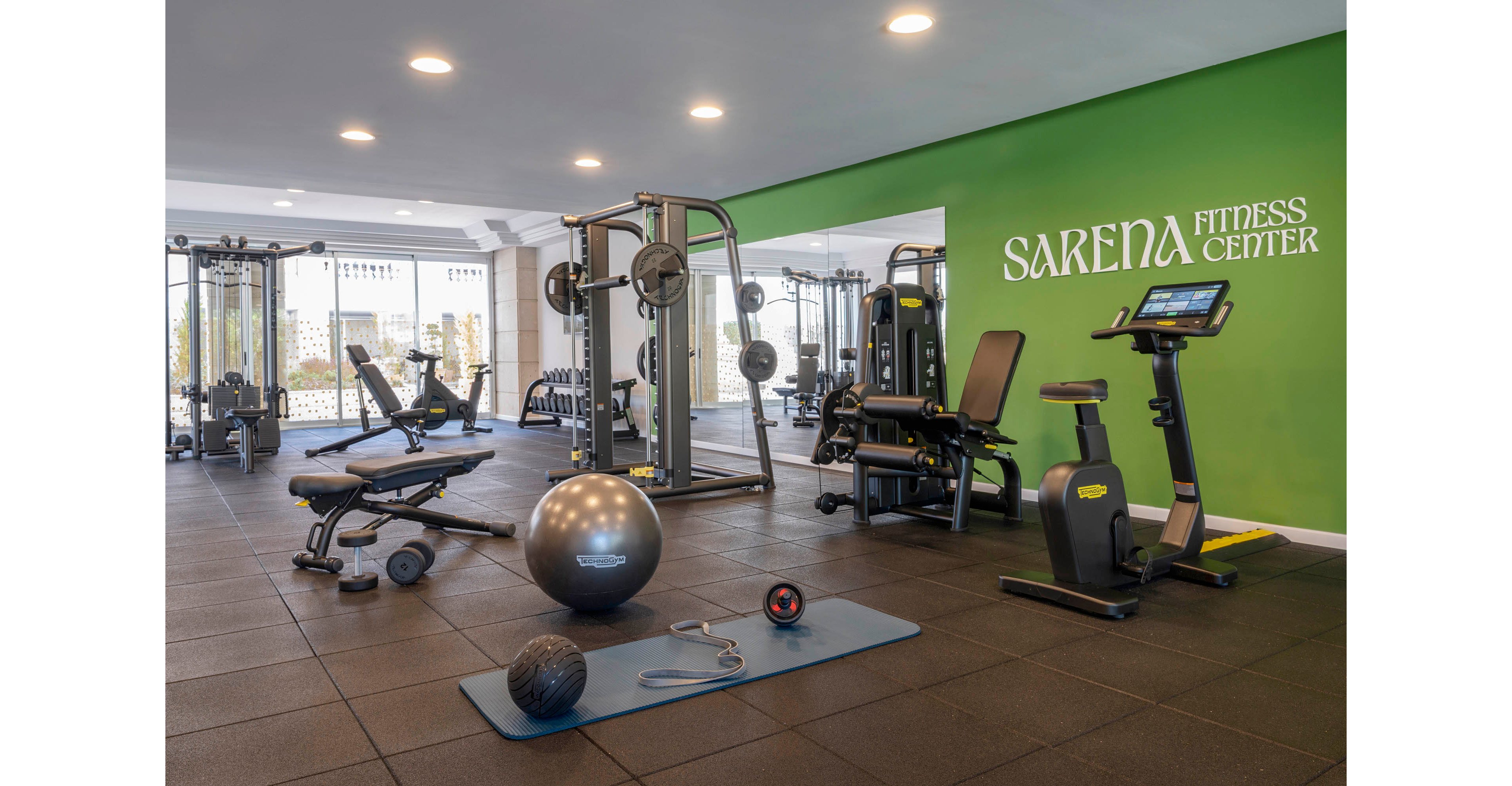 Spacious indoor gym featuring various strength and cardio machines. The Sarena Fitness Center name is displayed on a bright green accent wall. Free weights, benches, an exercise bike, stability ball, and other workout accessories are neatly arranged on rubber flooring. Large windows in the background let in natural light, creating an inviting workout environment.