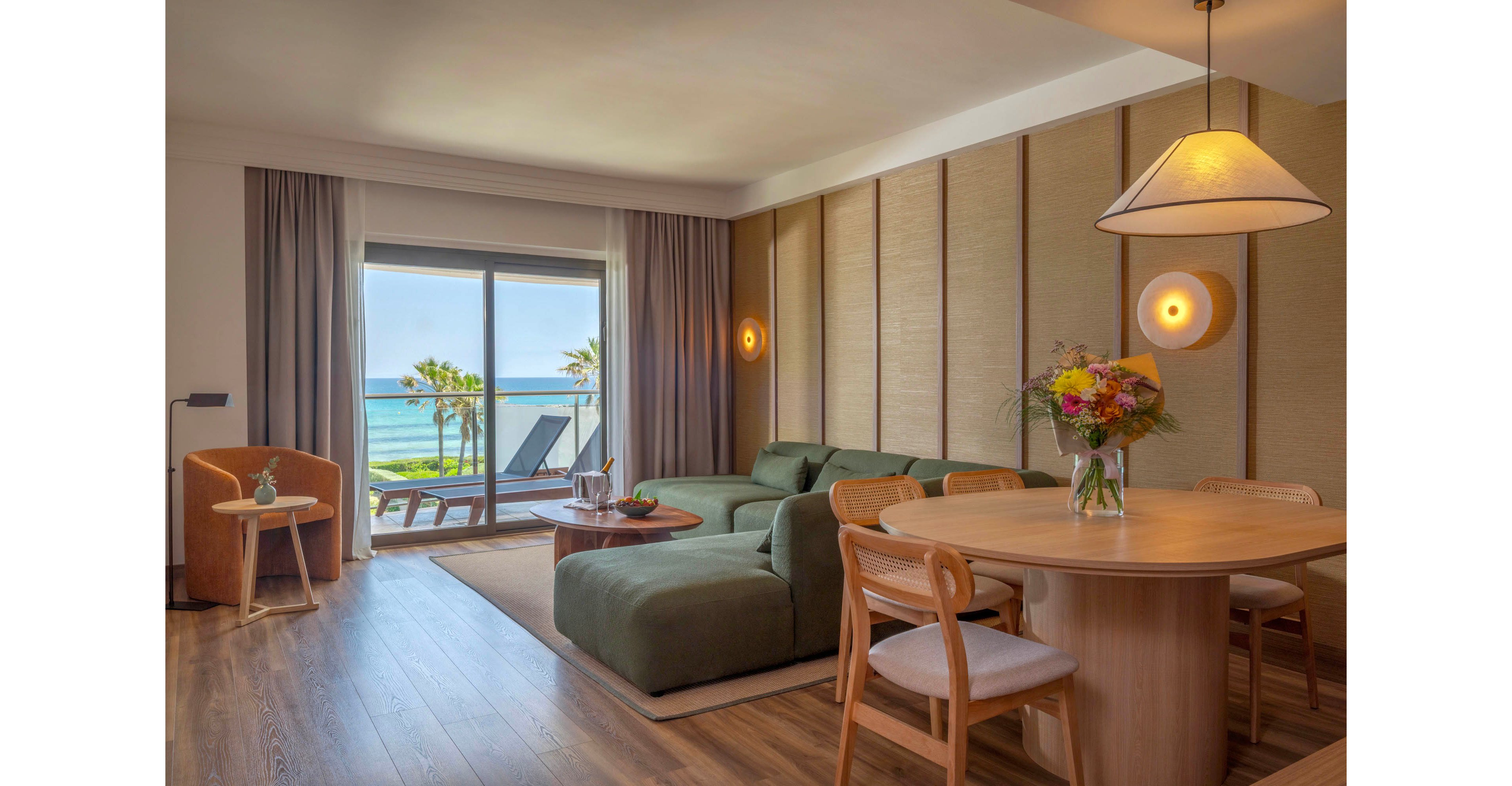 A contemporary hotel suite features a spacious living and dining area with a large green sectional sofa and round wooden table. Floor-to-ceiling glass doors open to a balcony overlooking palm trees and a calm ocean. Warm wall sconces, a pendant lamp, and neutral-toned curtains create a cozy, inviting atmosphere. Fresh flowers, fruit, and drinks on the tables add colorful accents to the serene coastal interior.