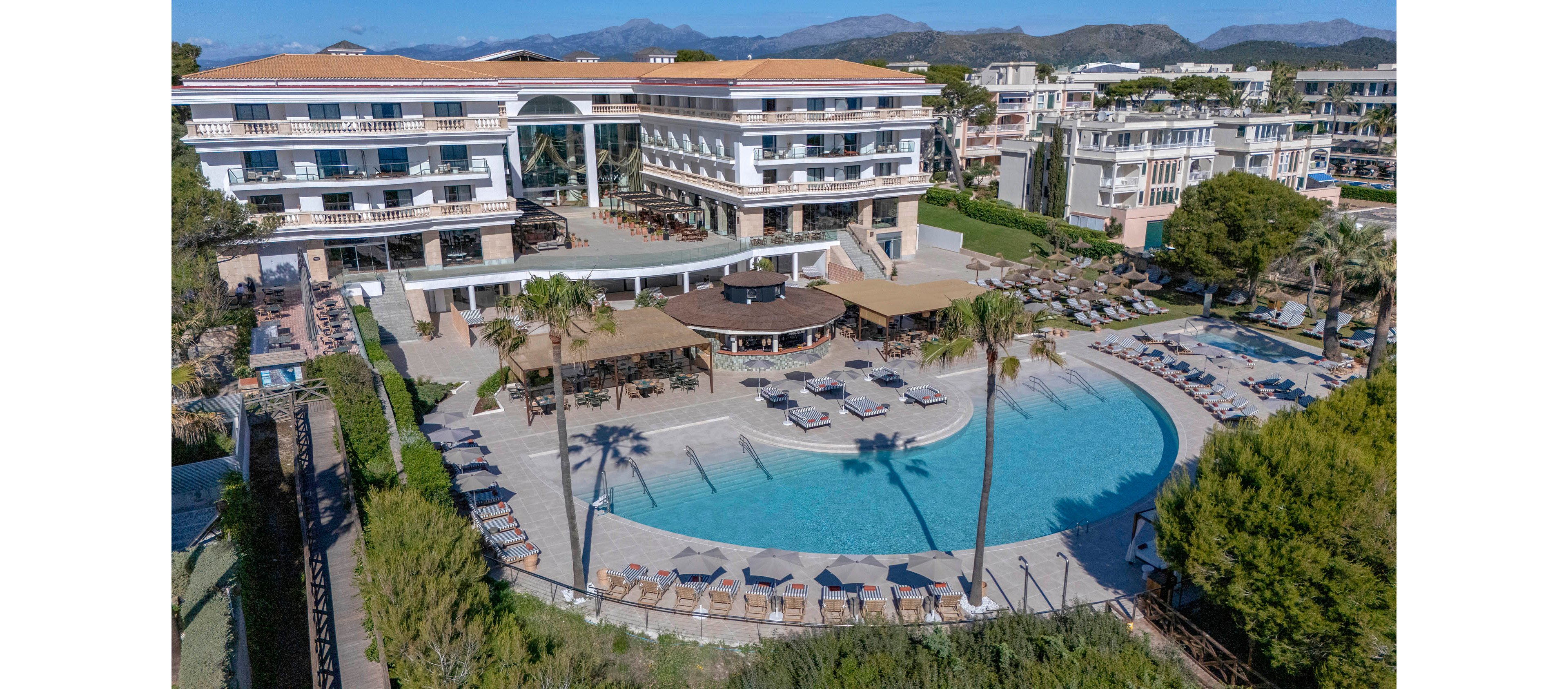 A large resort complex with multiple stories overlooks a spacious, curved outdoor swimming pool. Sun loungers and umbrellas are neatly arranged around the pool, creating a relaxing vacation atmosphere. Palm trees and landscaped greenery frame the pool deck, while additional buildings and mountains are visible in the background. The bright daylight and clear blue water emphasize a sunny, Mediterranean-style holiday setting.