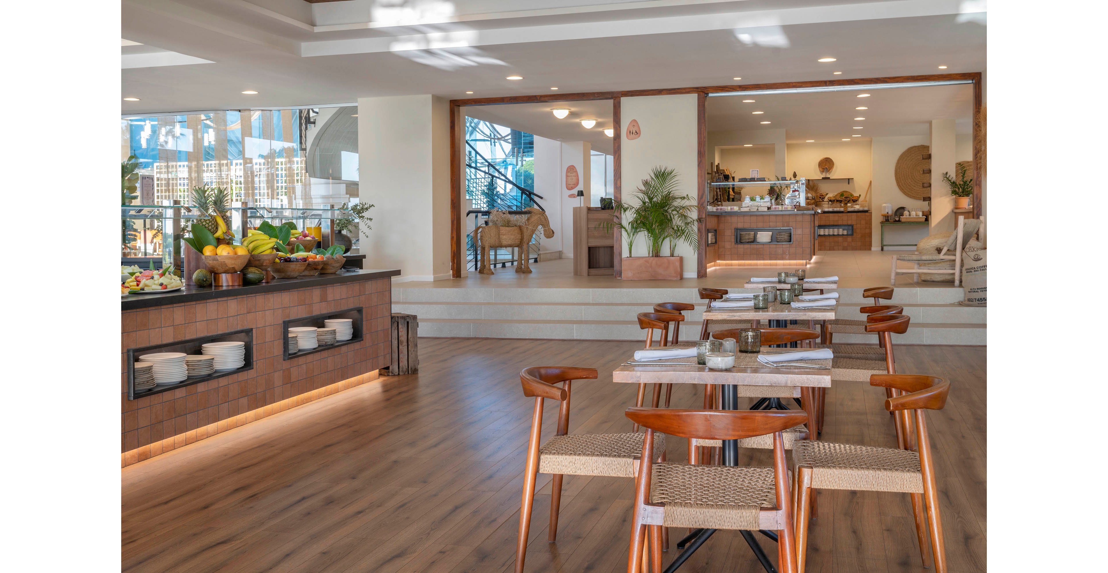 Bright contemporary restaurant interior featuring wooden tables and woven chairs is shown. A buffet counter on the left displays fresh fruits, vegetables, and breakfast items. The open-plan space leads to a service area with additional food stations and decor. Large windows and warm lighting create an inviting, relaxed dining atmosphere.