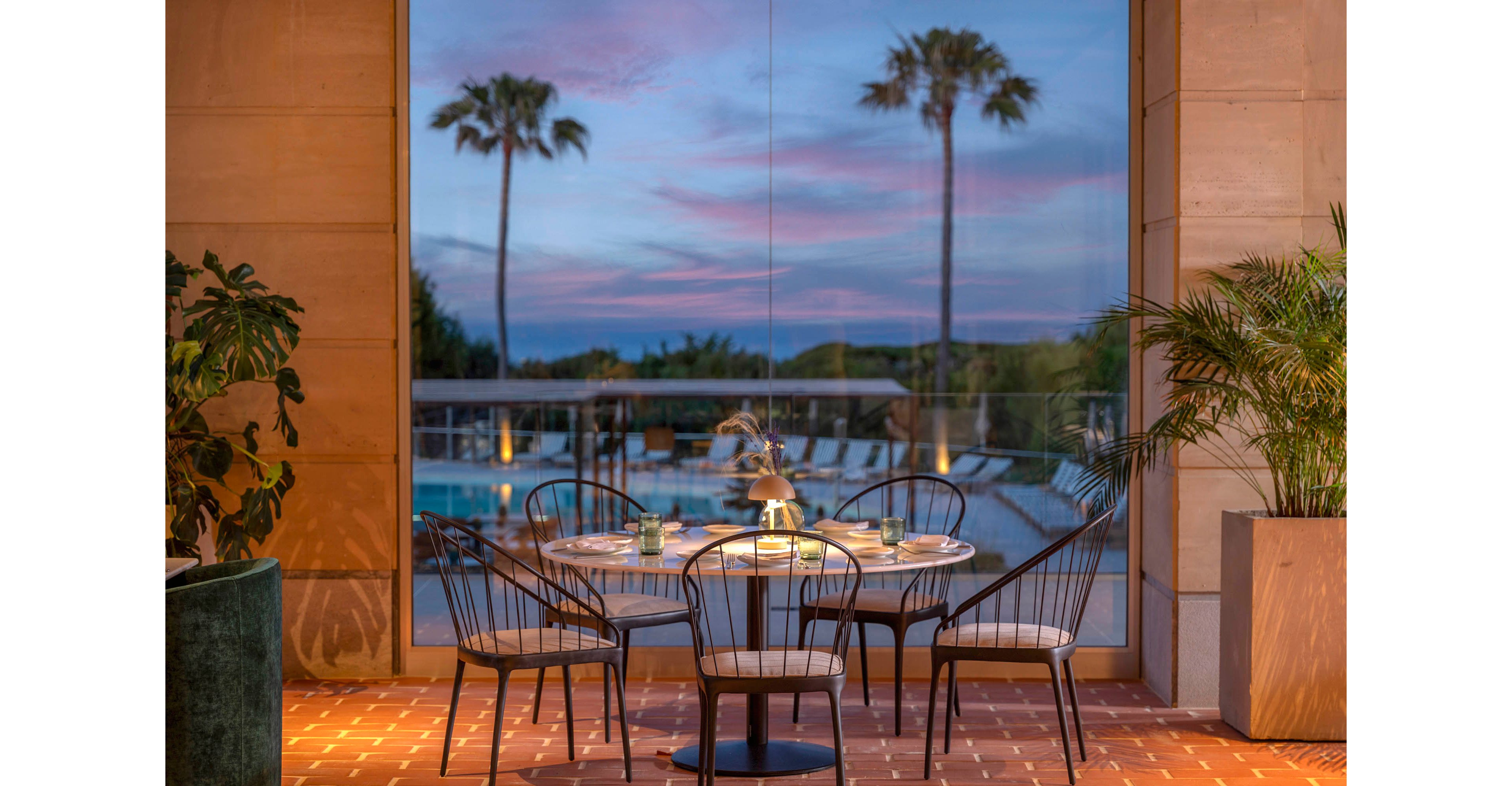 An elegant outdoor dining table is set for four on a tiled terrace. The scene overlooks a swimming pool framed by tall palm trees at dusk. Soft lighting from a table lamp and surrounding fixtures creates a warm, inviting atmosphere. Potted plants and modern metal chairs enhance the relaxed, upscale resort setting.