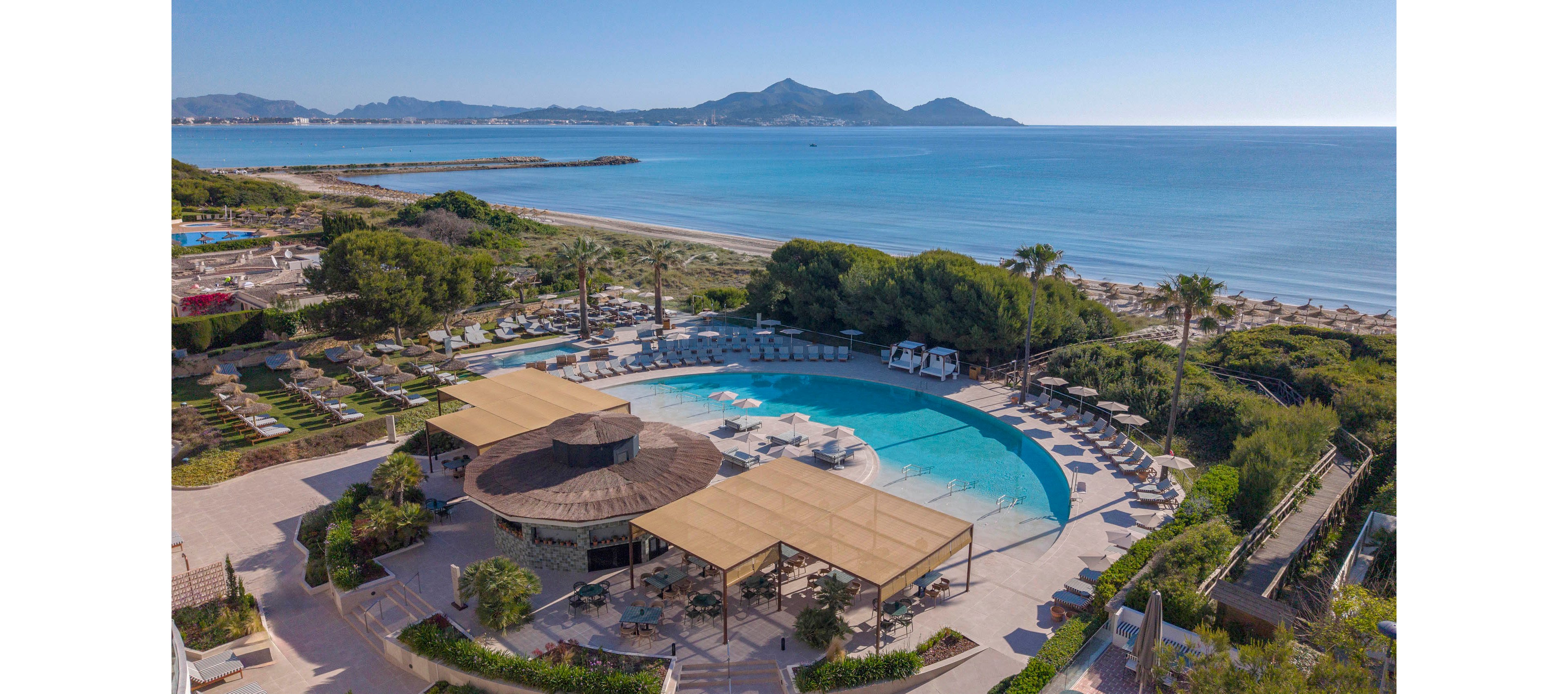 A large circular swimming pool sits at the center of a coastal resort. Sun loungers and umbrellas surround the pool, with landscaped gardens and pathways leading toward the beach. The sandy shoreline stretches along a calm blue sea, with gentle waves meeting the coast. Distant mountains frame the horizon under a clear, sunny sky, creating a tranquil vacation atmosphere.