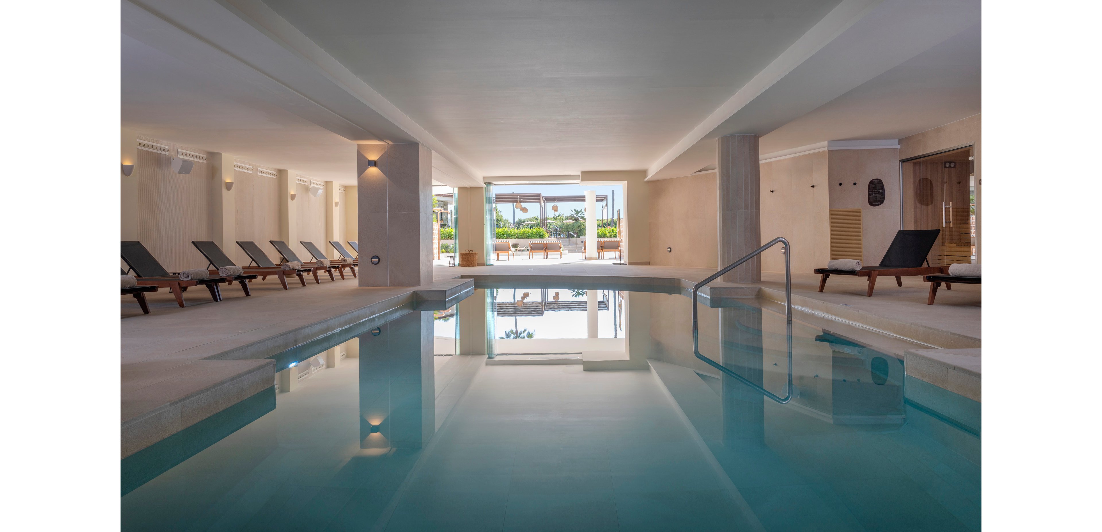 Calm indoor swimming pool area with clear blue water and modern minimalist architecture. Dark cushioned loungers line both sides of the pool, inviting guests to relax. Soft wall lighting and neutral beige tones create a tranquil spa-like atmosphere. Large openings at the far end reveal an outdoor terrace and greenery, bringing natural light into the space.