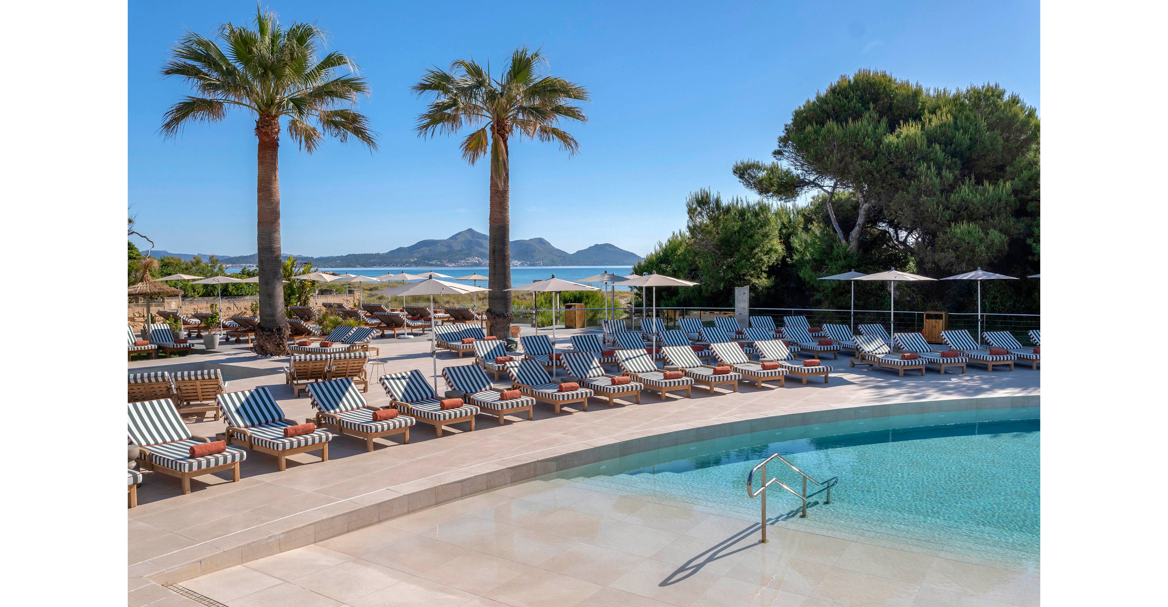 An outdoor resort swimming pool is surrounded by neatly arranged striped sun loungers and umbrellas. Tall palm trees and lush greenery frame the pool area, creating a relaxing vacation atmosphere. In the background, a sandy beach and calm blue sea stretch toward distant mountains under a clear sky. The scene suggests a tranquil, sunny day at a coastal holiday destination.