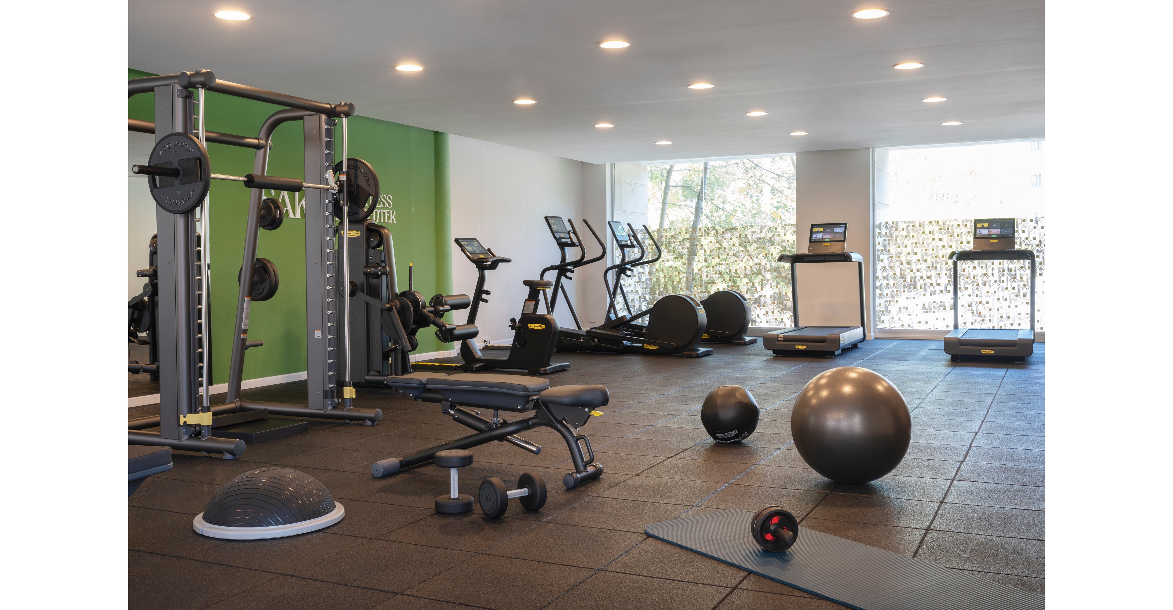 Spacious modern gym interior featuring various strength and cardio equipment. A weight machine, adjustable bench, dumbbells, and balance trainer are arranged on rubber flooring in the foreground. Treadmills, stationary bikes, and large exercise balls are visible toward the back of the room. Recessed ceiling lights and large windows create a bright, inviting workout environment.