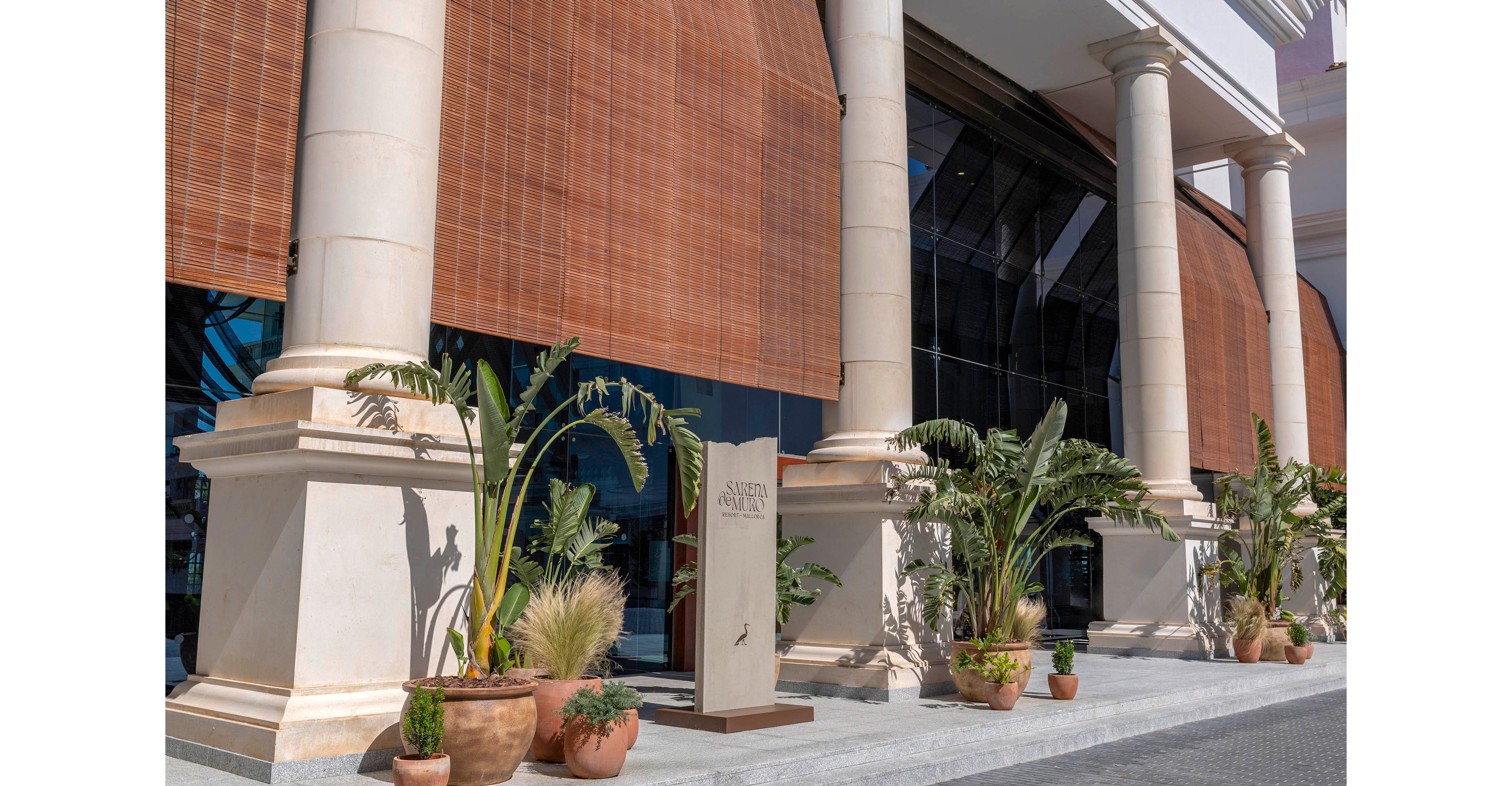 A grand building entrance features tall stone columns and large bamboo-style blinds covering the glass facade. Several potted plants and palms are arranged along the walkway, adding greenery to the architectural scene. A vertical sign stands near the entrance, partially readable but indicating the name of the establishment. Bright daylight and clear shadows suggest a warm, sunny climate and an upscale, resort-like atmosphere.