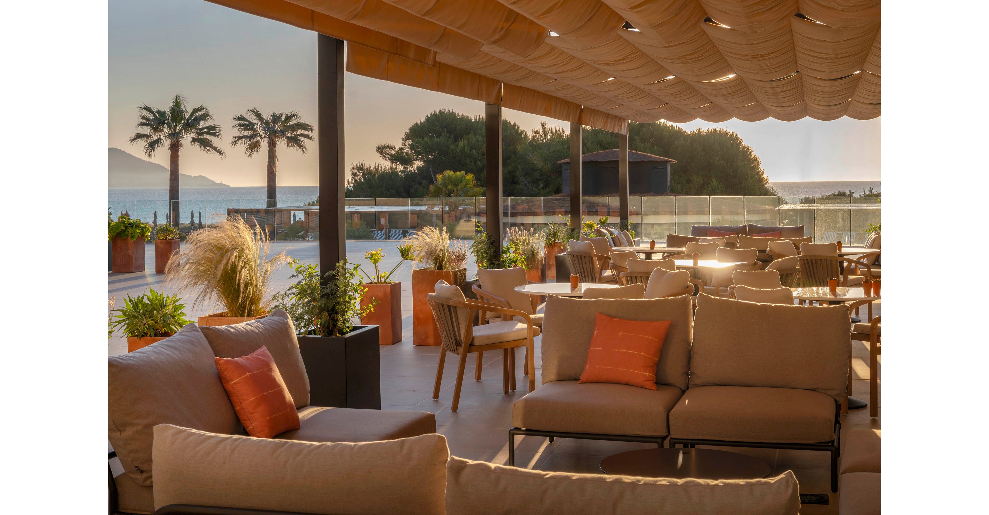 An open-air terrace lounge overlooks the sea at sunset, furnished with cushioned sofas and dining tables. Warm sunlight filters through the canopy roof, casting soft shadows across the seating area. Potted plants and ornamental grasses add greenery and texture along the edges of the space. In the background, palm trees, distant hills, and calm water create a relaxed coastal atmosphere.