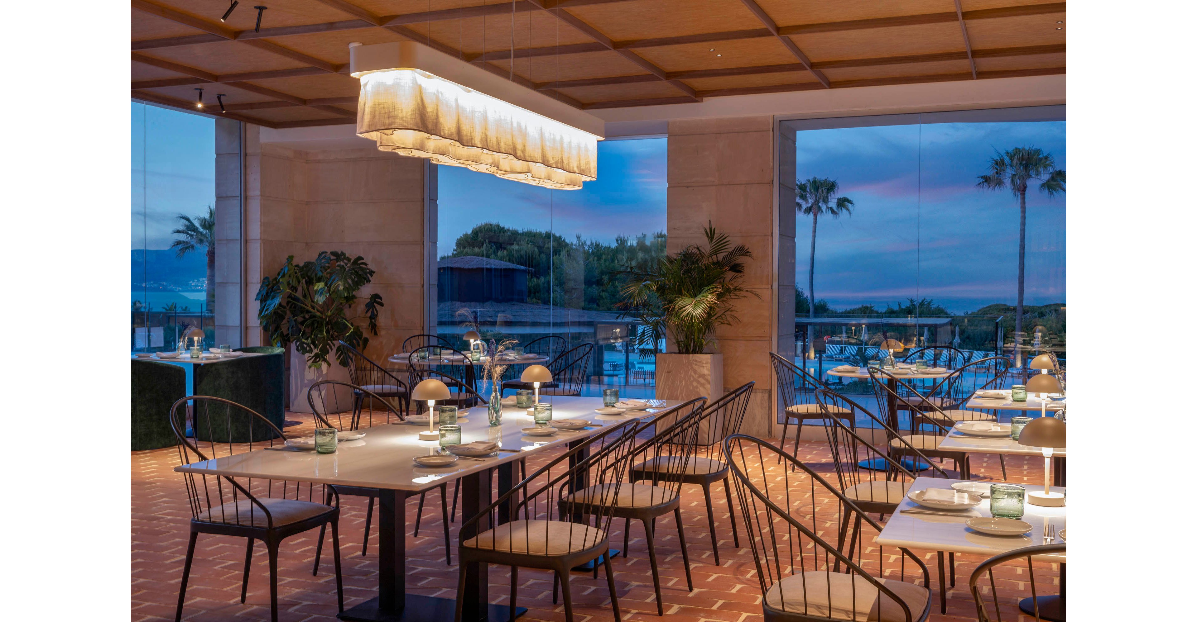 Contemporary restaurant tables are set for dinner inside a bright, glass-walled dining room. A large rectangular pendant light hangs from a wooden ceiling, illuminating neatly arranged chairs and table settings. Floor-to-ceiling windows reveal an outdoor terrace with palm trees and a colorful evening sky over the water. Potted plants and warm lighting create a relaxed yet upscale coastal ambiance.