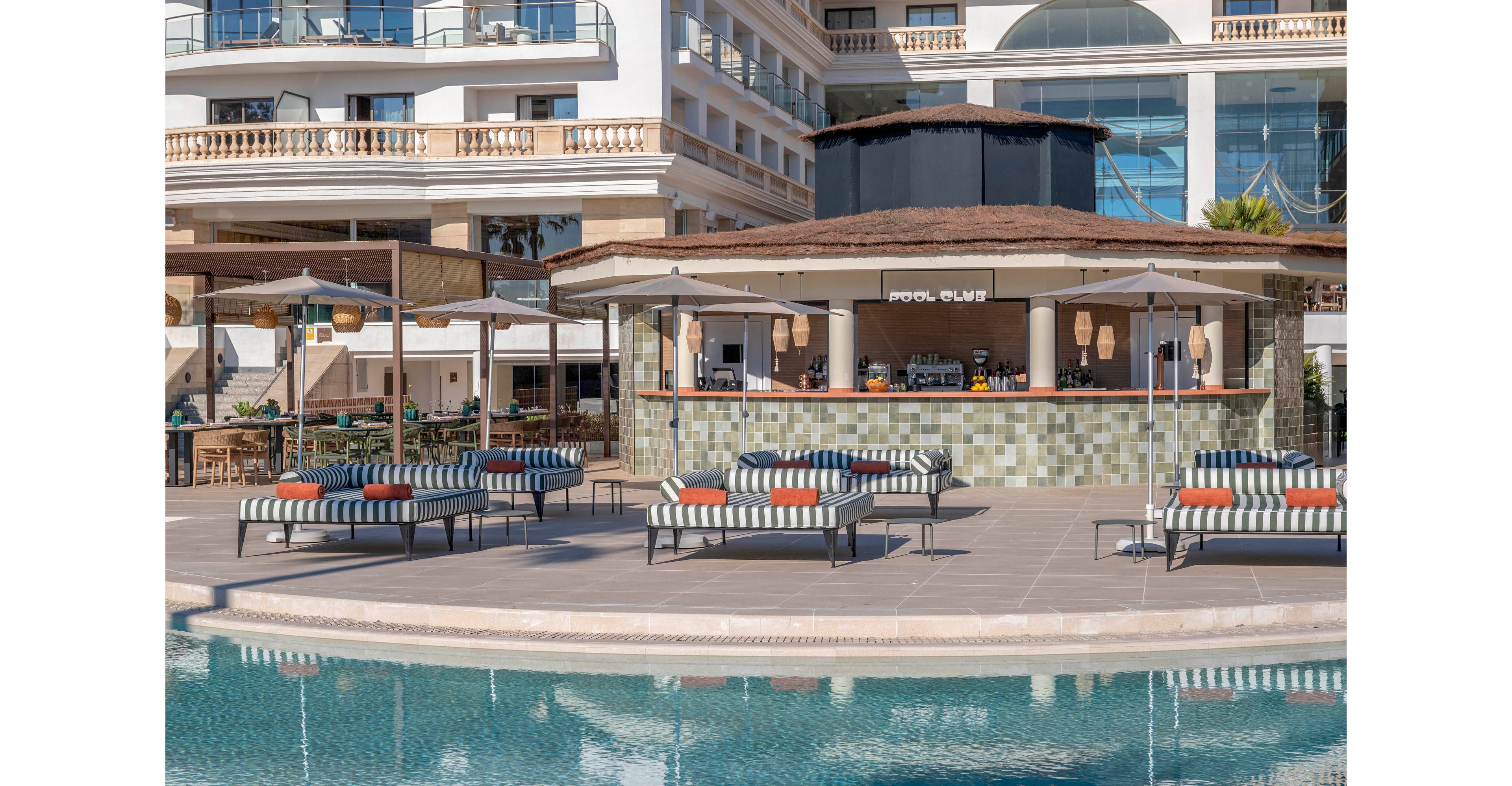 An outdoor resort pool area features a central bar labeled "POOL CLUB" facing the water. Striped cushioned lounge chairs and umbrellas are arranged neatly around the pool deck, creating an inviting relaxation space. The modern hotel building with balconies and large windows rises in the background. The scene is calm and sunny, suggesting a leisurely vacation atmosphere.