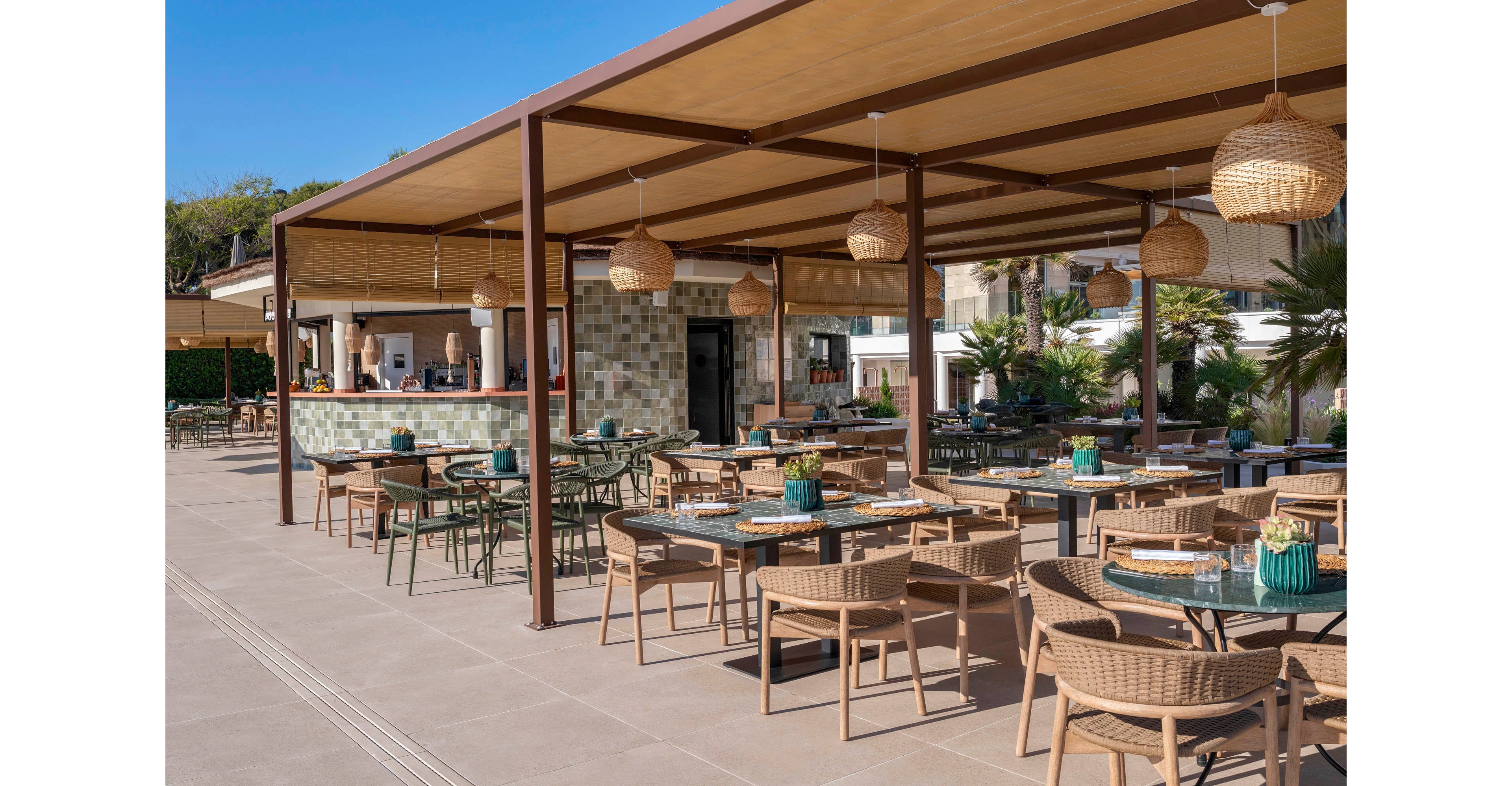 An open-air terrace restaurant features neatly arranged tables and wicker chairs under a large wooden pergola. A bar area with tiled front and barstools sits to the side, ready for guests. Woven pendant lights hang from the ceiling, adding a warm, relaxed ambiance. In the background, palm trees and resort buildings suggest a sunny vacation destination.