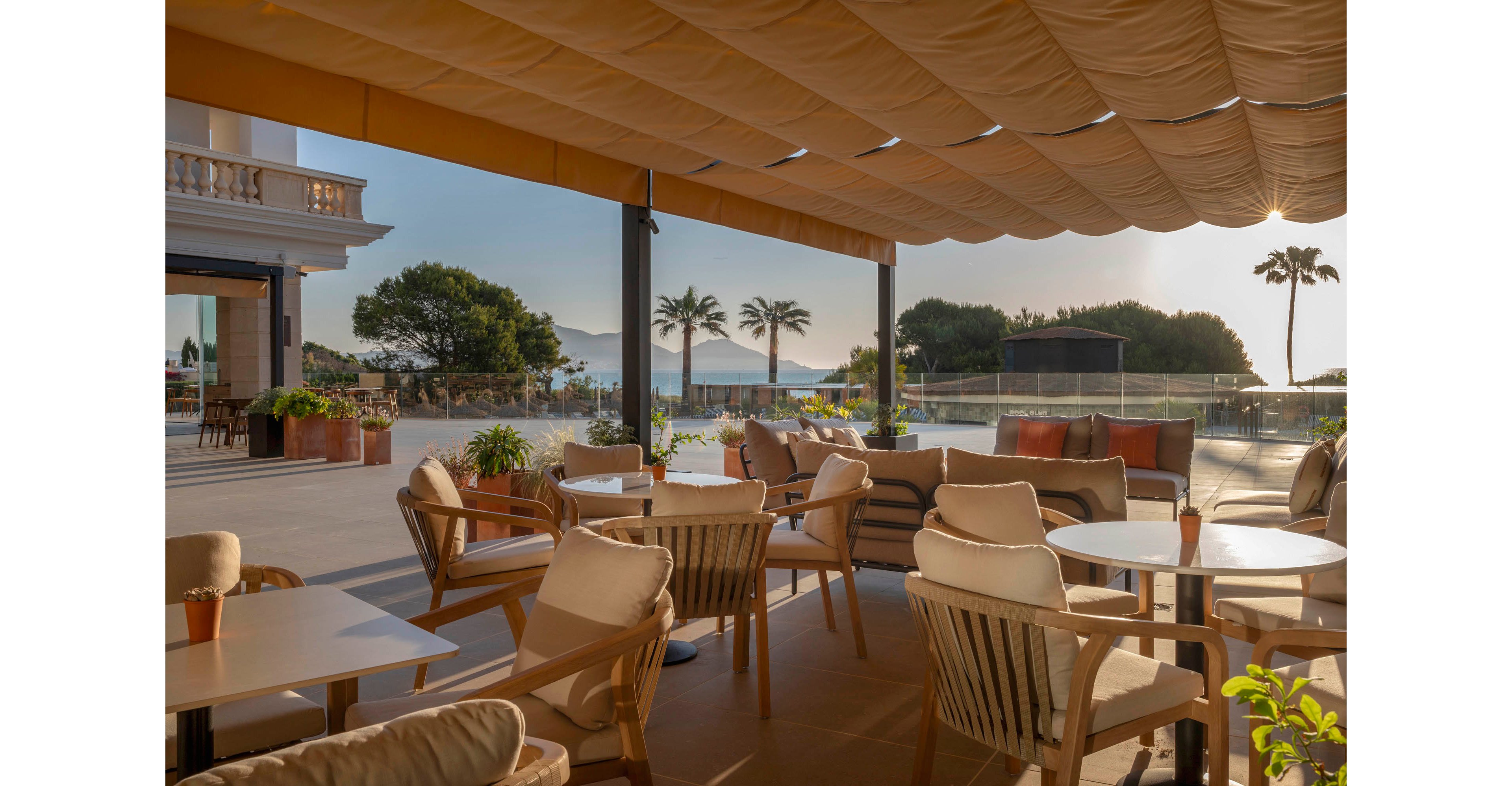 An inviting outdoor terrace features cushioned wooden chairs and round tables arranged under a large fabric canopy. Warm sunlight casts soft shadows across the tiled floor, creating a relaxed atmosphere. In the background, palm trees and distant mountains frame a calm body of water. The scene suggests a tranquil coastal resort or café setting ideal for leisurely dining or drinks.