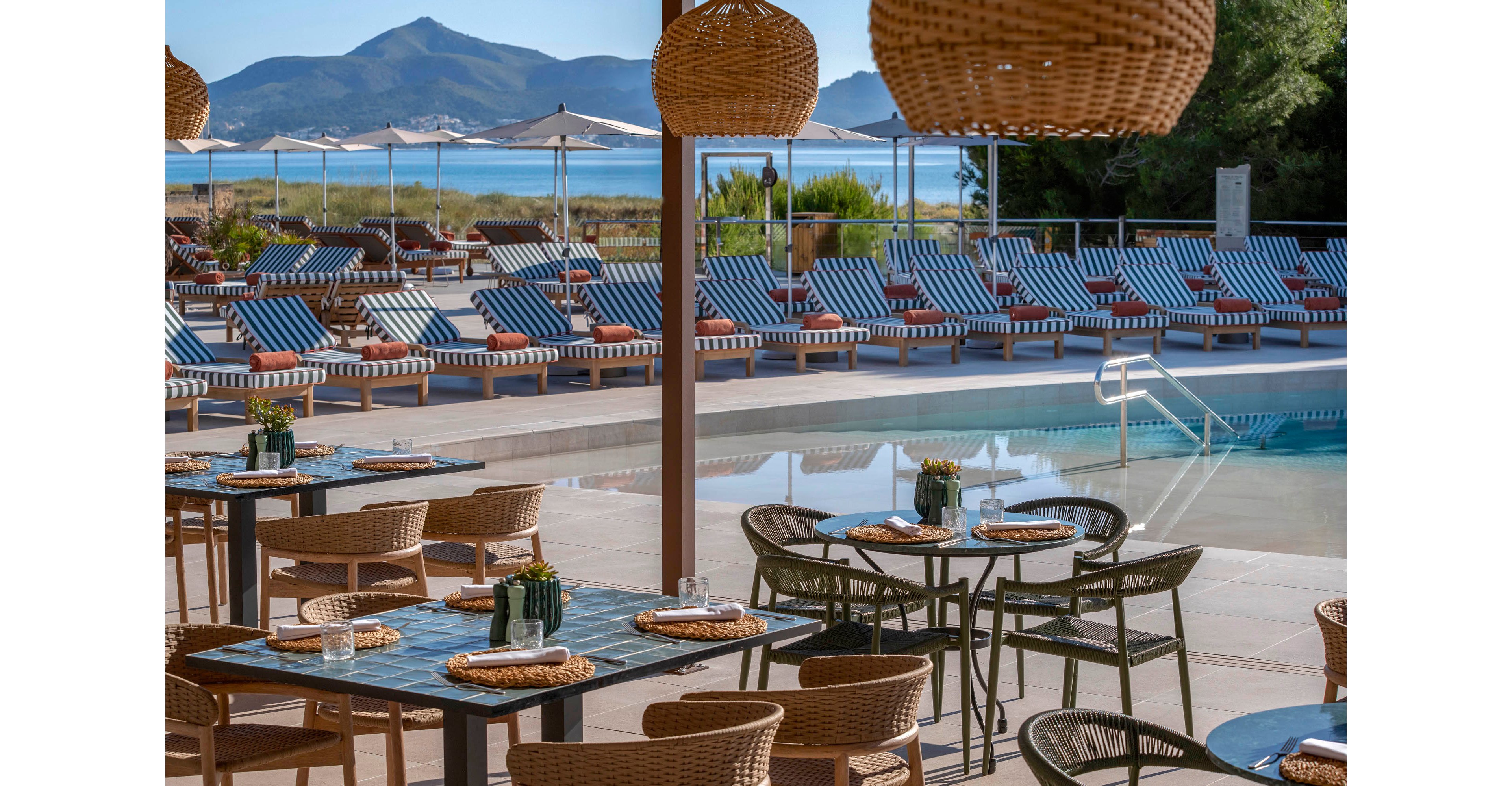 An outdoor resort dining area overlooks a swimming pool lined with striped sun loungers. Wicker chairs and hanging woven lamps create a relaxed, coastal atmosphere under a shaded terrace. Small vases with flowers decorate the tables, which are set with placemats and glassware. In the background, calm water and distant mountains complete the scenic vacation setting.