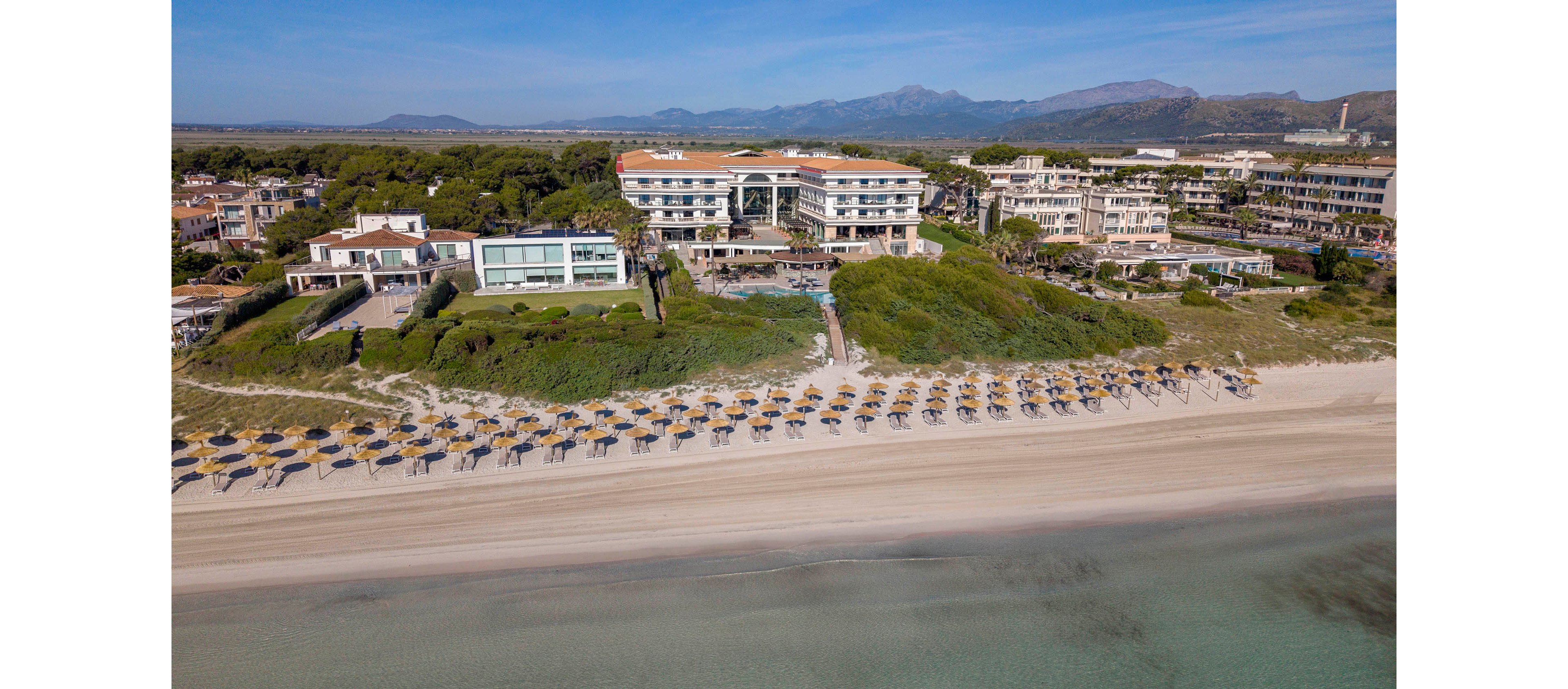 A large beachfront resort complex is captured from above along a wide sandy shoreline. Rows of sun loungers and umbrellas line the beach facing calm, shallow water. Behind the resort, low-rise buildings and green vegetation extend toward distant mountains under a clear blue sky. The scene suggests a tranquil holiday destination with easy access to the sea and scenic natural surroundings.