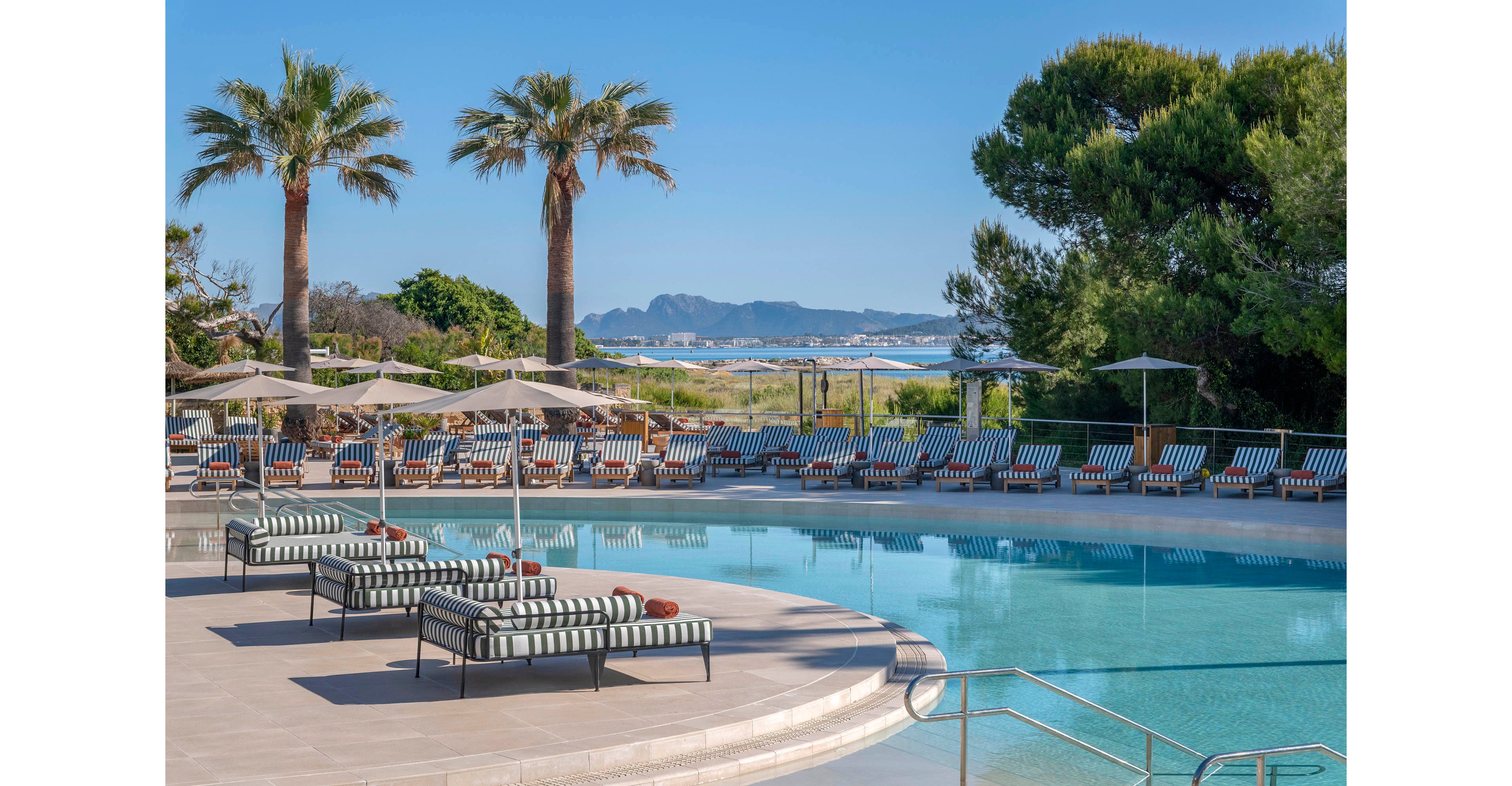 A tranquil resort swimming pool is surrounded by striped cushioned sun loungers and parasols. Tall palm trees and lush greenery frame the pool area, creating a relaxing vacation atmosphere. In the distance, a sandy shoreline and calm blue sea lead to hazy mountains under a clear sky. The scene is bright and sunny, suggesting a warm coastal destination ideal for leisure and sunbathing.