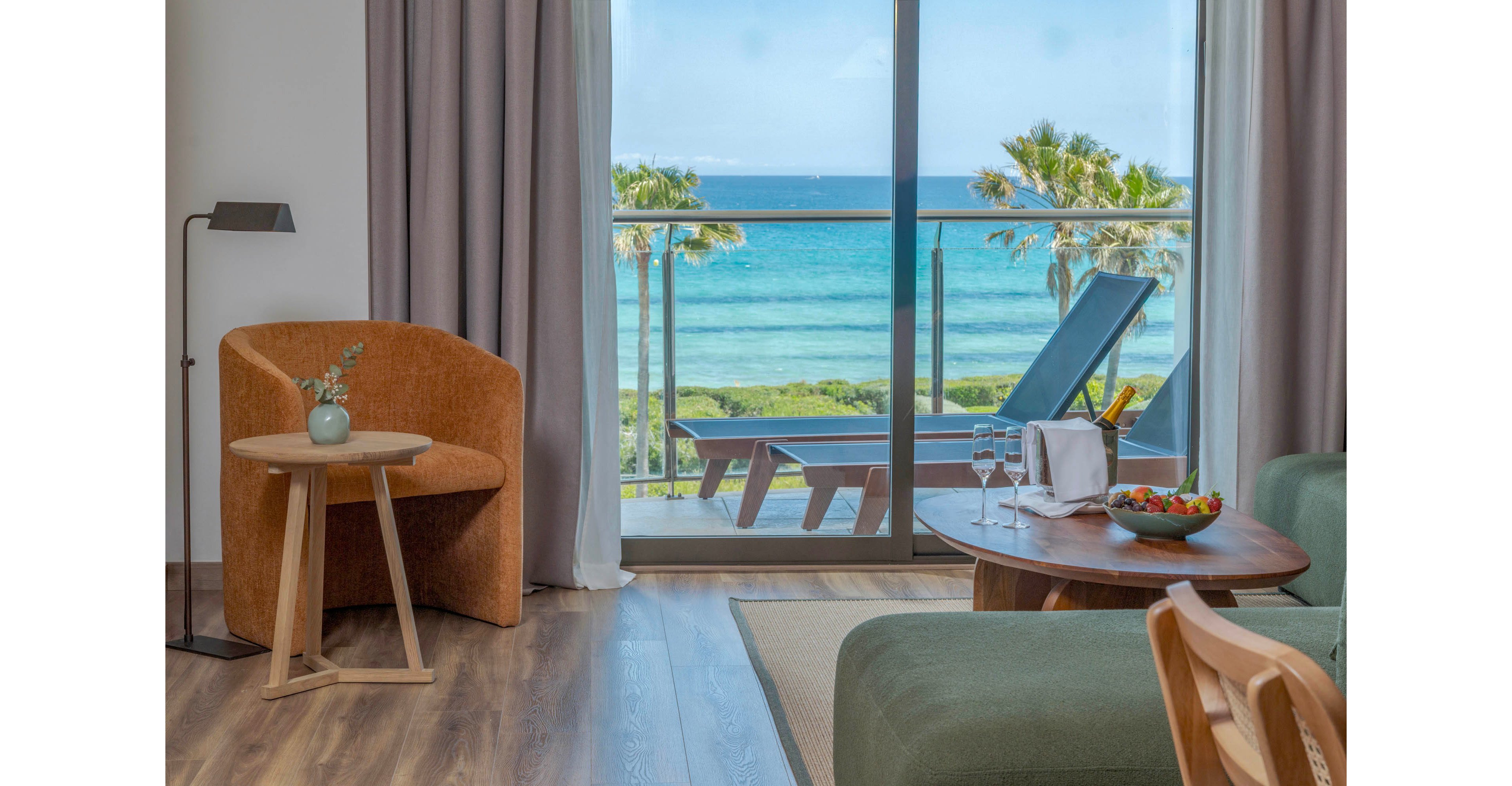 A contemporary hotel room opens onto a balcony overlooking a turquoise ocean and palm trees. Inside, a cozy armchair and small side table sit beside floor-to-ceiling curtains. A wooden coffee table holds two wine glasses, a bottle in a cooler, and a bowl of fresh fruit. On the balcony, two lounge chairs face the sea, creating a relaxing vacation atmosphere.