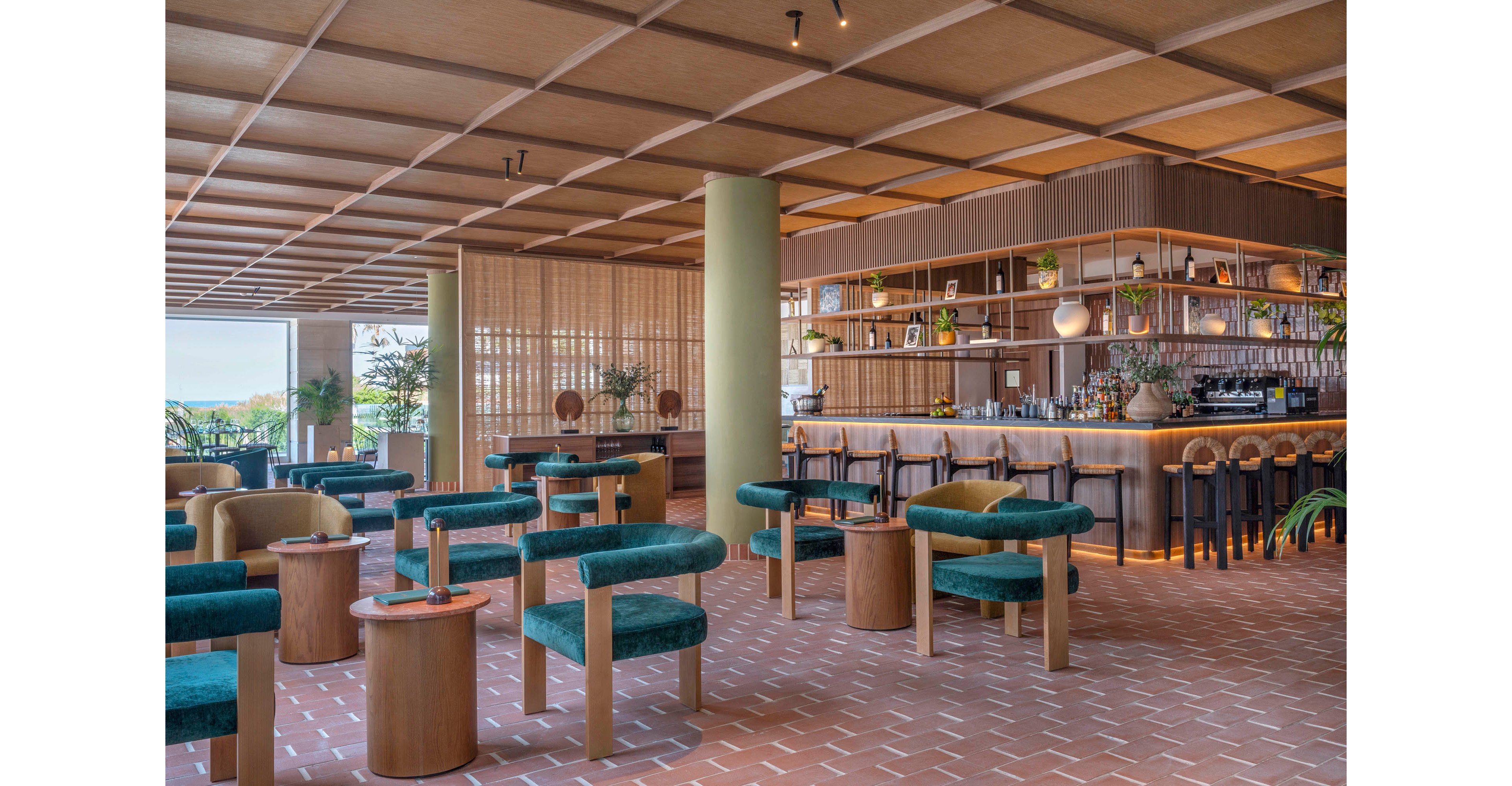 Contemporary lounge bar interior featuring low green upholstered chairs and round wooden tables. The space includes a long bar counter with high stools and a well-organized back bar of bottles and glassware. Warm wood tones, terracotta floor tiles, and soft ambient lighting create a relaxed, upscale atmosphere. Large windows and indoor plants add natural light and a fresh, airy feel to the setting.