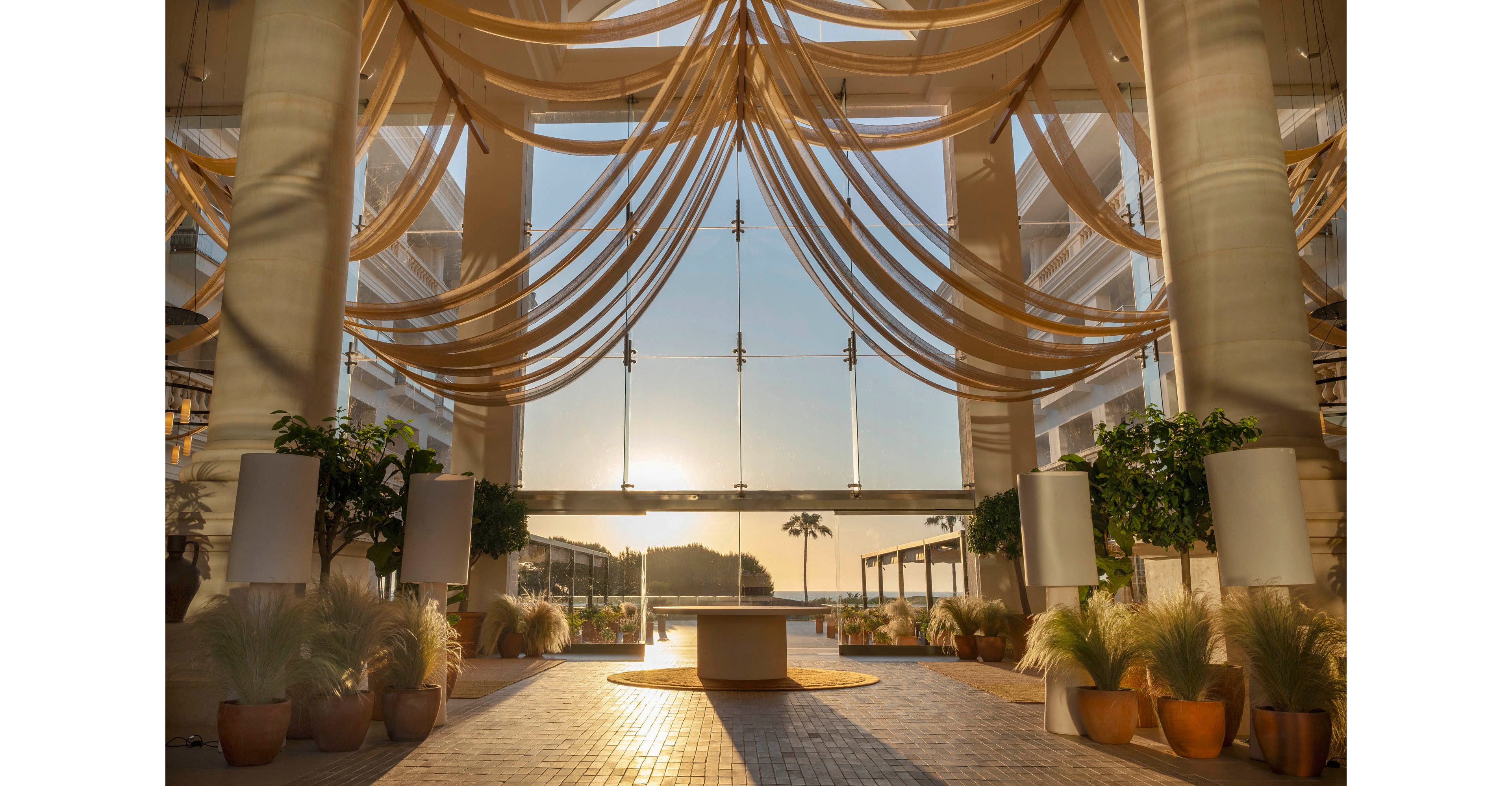 Spacious hotel lobby interior opens toward a scenic seaside view at sunset. Sheer fabric drapes hang in soft curves from the high ceiling, framing large glass windows and doors. Potted plants and ornamental grasses line the tiled walkway, adding lush greenery to the warm, golden space. The low sun glows over distant hills and palm trees, creating a tranquil, luxurious resort atmosphere.