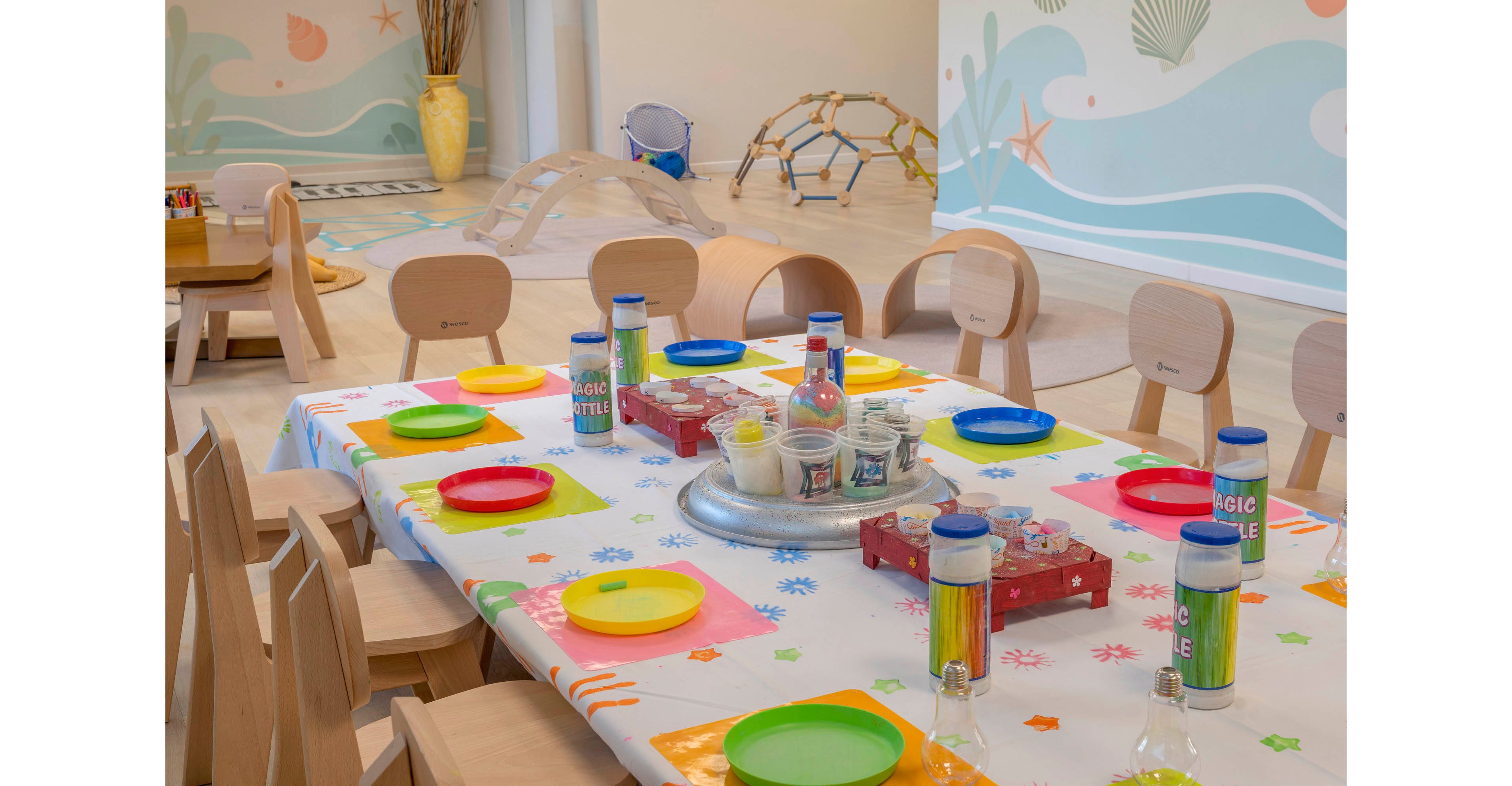 A long table is set for a children's party with bright plastic plates and cups arranged on a patterned tablecloth. Art supplies and bottles labeled "MAGIC BUBBLES" sit in the center of the table. Wooden chairs surround the table, and additional small tables and chairs are visible in the background. The playroom features light wooden floors, soft pastel wall murals with sea-themed illustrations, and various wooden play structures.