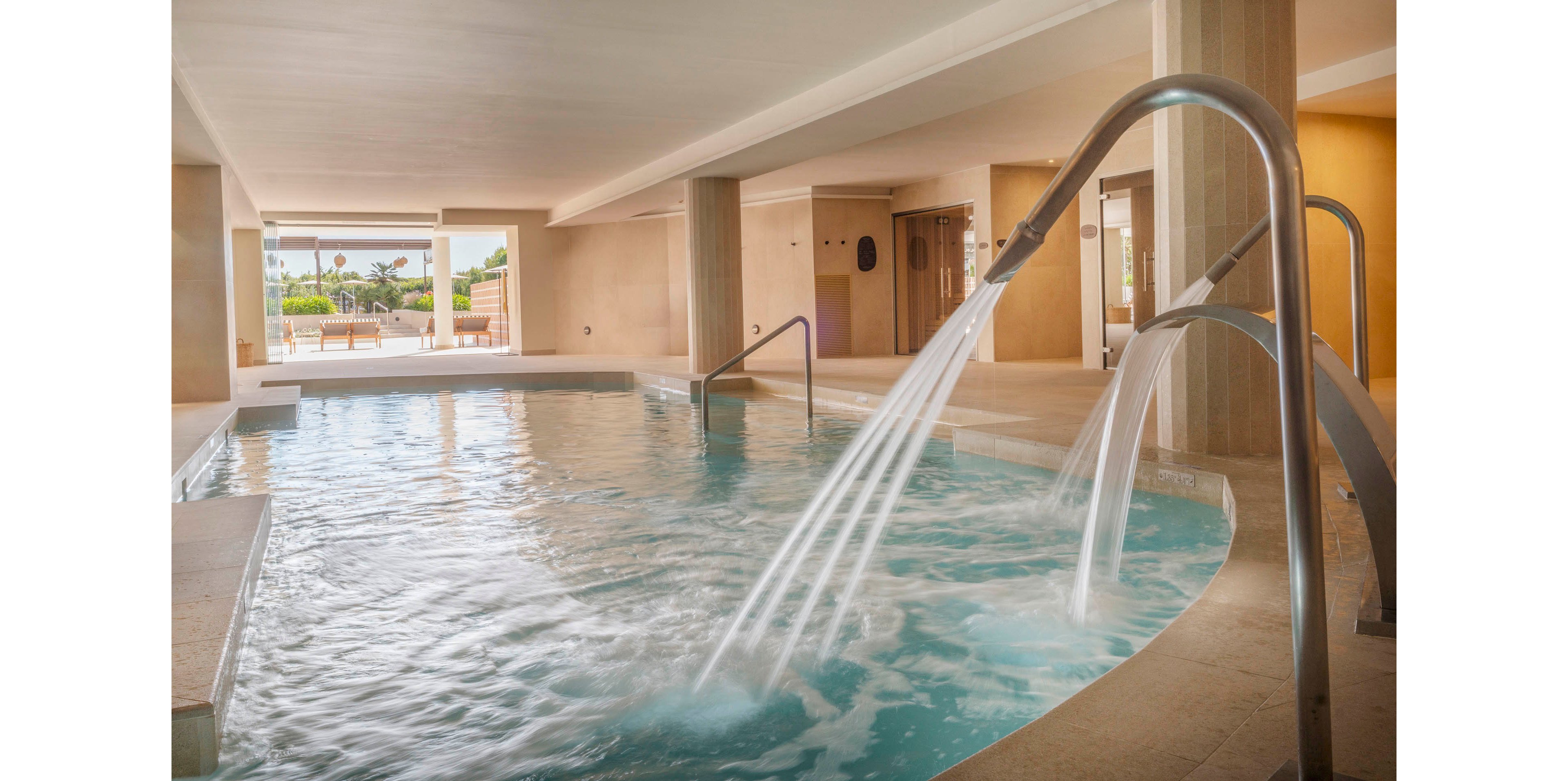 Indoor spa pool area featuring curved metal water jets spraying into turquoise water. The space is surrounded by beige stone columns and walls, creating a warm, relaxing atmosphere. Natural light enters from large openings leading to an outdoor terrace with greenery and palm trees. The scene suggests a tranquil wellness or hotel spa environment designed for relaxation and hydrotherapy.