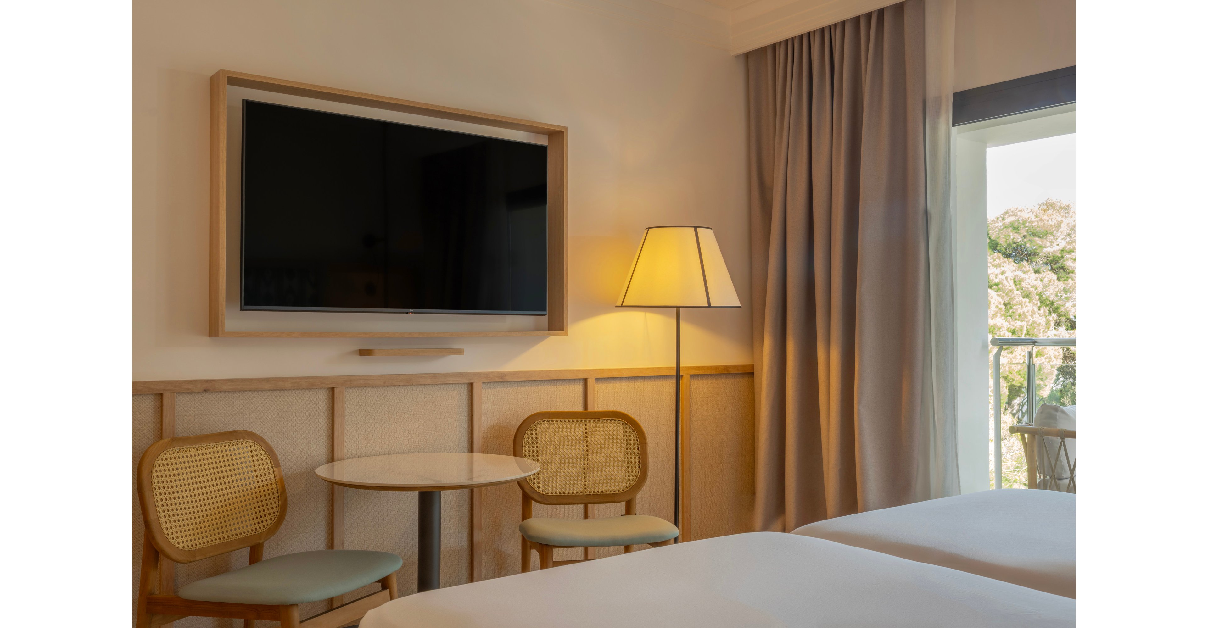 A contemporary hotel room features a large flat-screen television mounted on the wall above a light wood panel. Two woven-back chairs and a small round table create a cozy seating area beside a tall floor lamp. Soft gray curtains frame a glass door that opens to a balcony with greenery visible outside. The neutral color palette and natural light give the space a calm, inviting atmosphere.