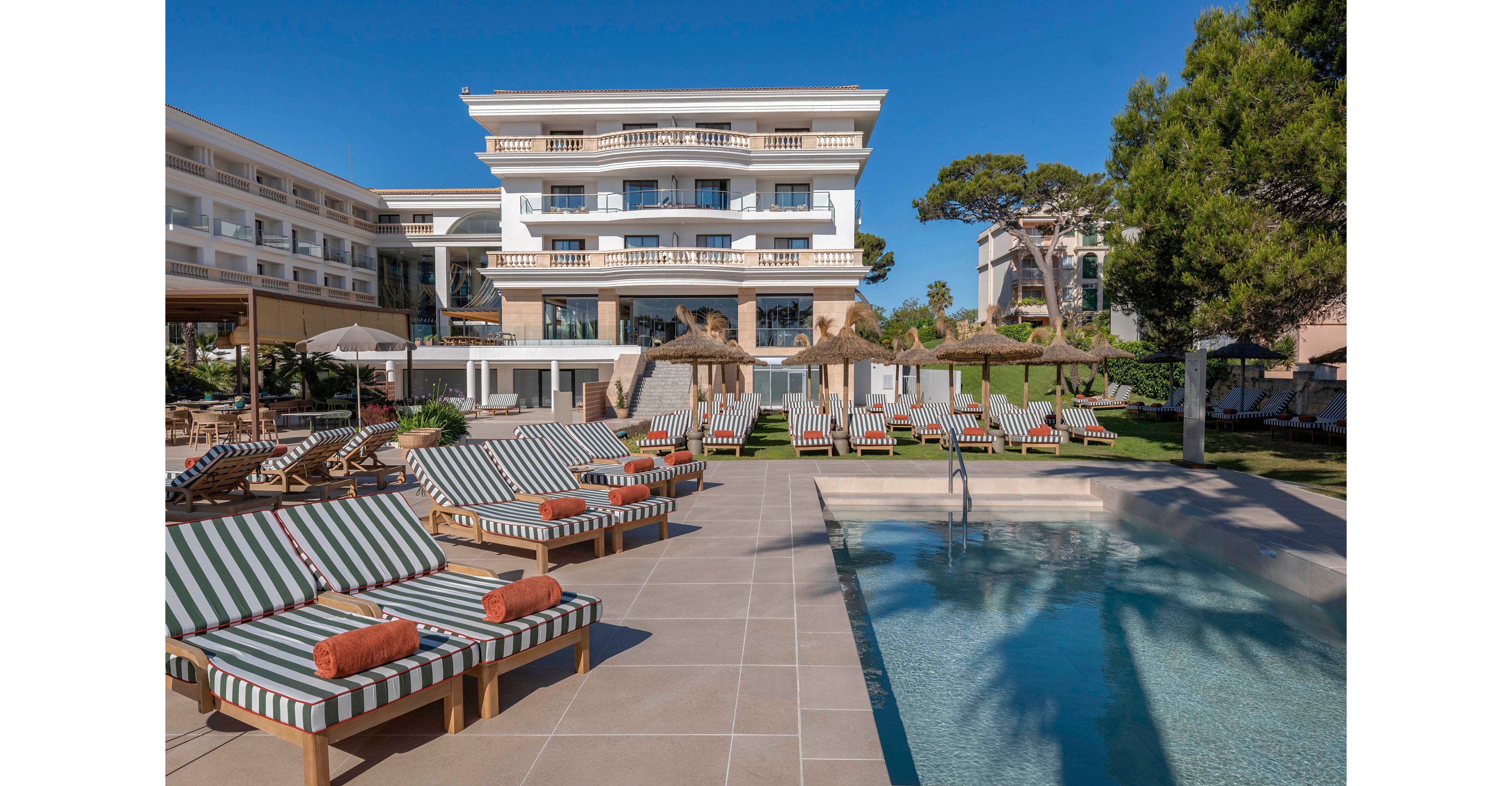 An upscale resort features a row of striped cushioned loungers arranged beside a clear outdoor swimming pool. The multi-story hotel building with balconies stands in the background, surrounded by trees and manicured lawns. Straw parasols and neatly rolled orange towels add to the sense of comfort and relaxation. The bright daylight and blue sky create a warm, inviting vacation atmosphere.