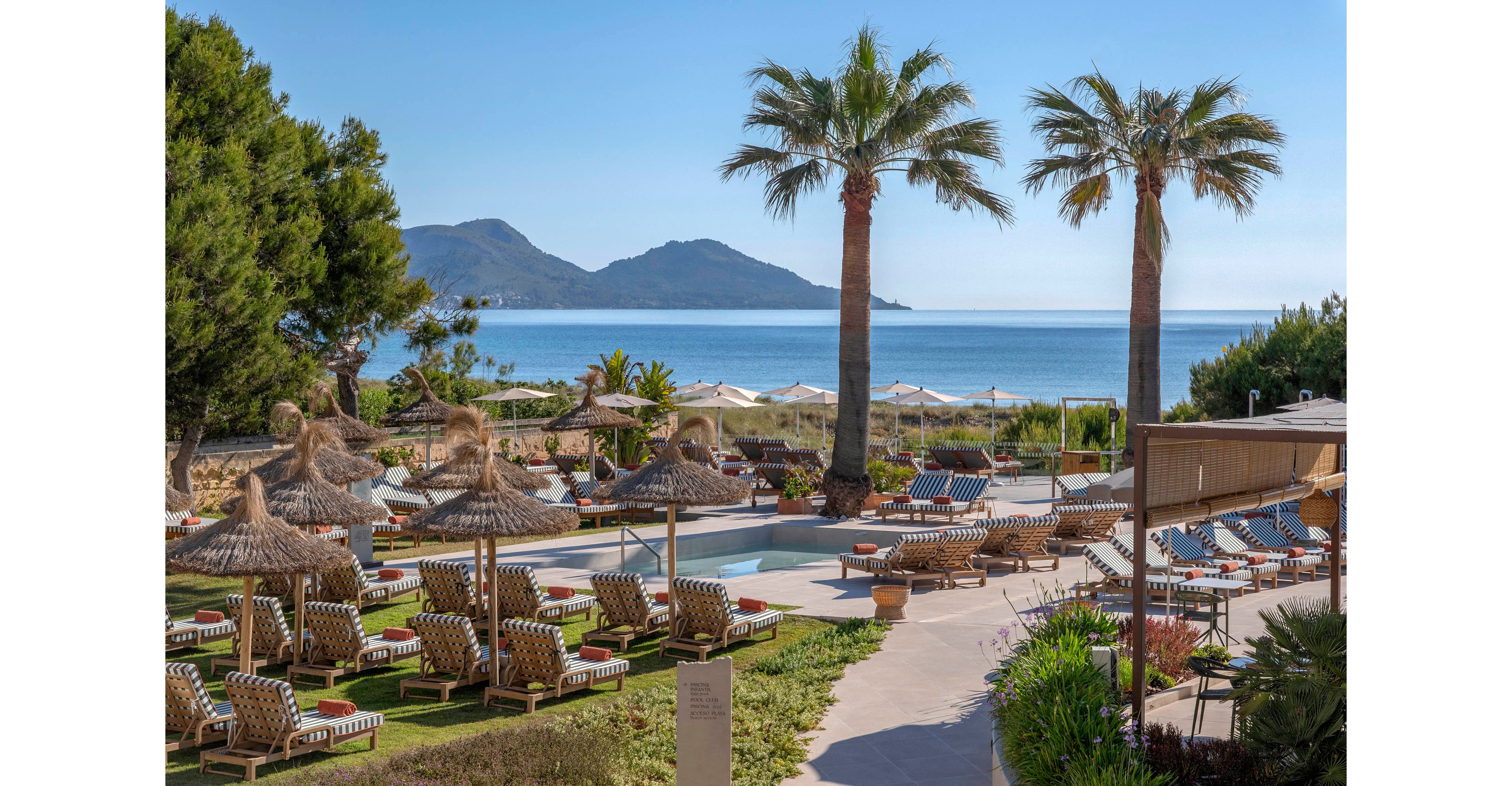 A coastal resort features a central swimming pool surrounded by sun loungers and straw parasols. Tall palm trees frame the scene, with manicured lawns and pathways leading toward the water. In the background, a calm blue sea stretches out to distant mountains under a clear sky. The setting suggests a relaxing vacation atmosphere at a beachfront hotel.
