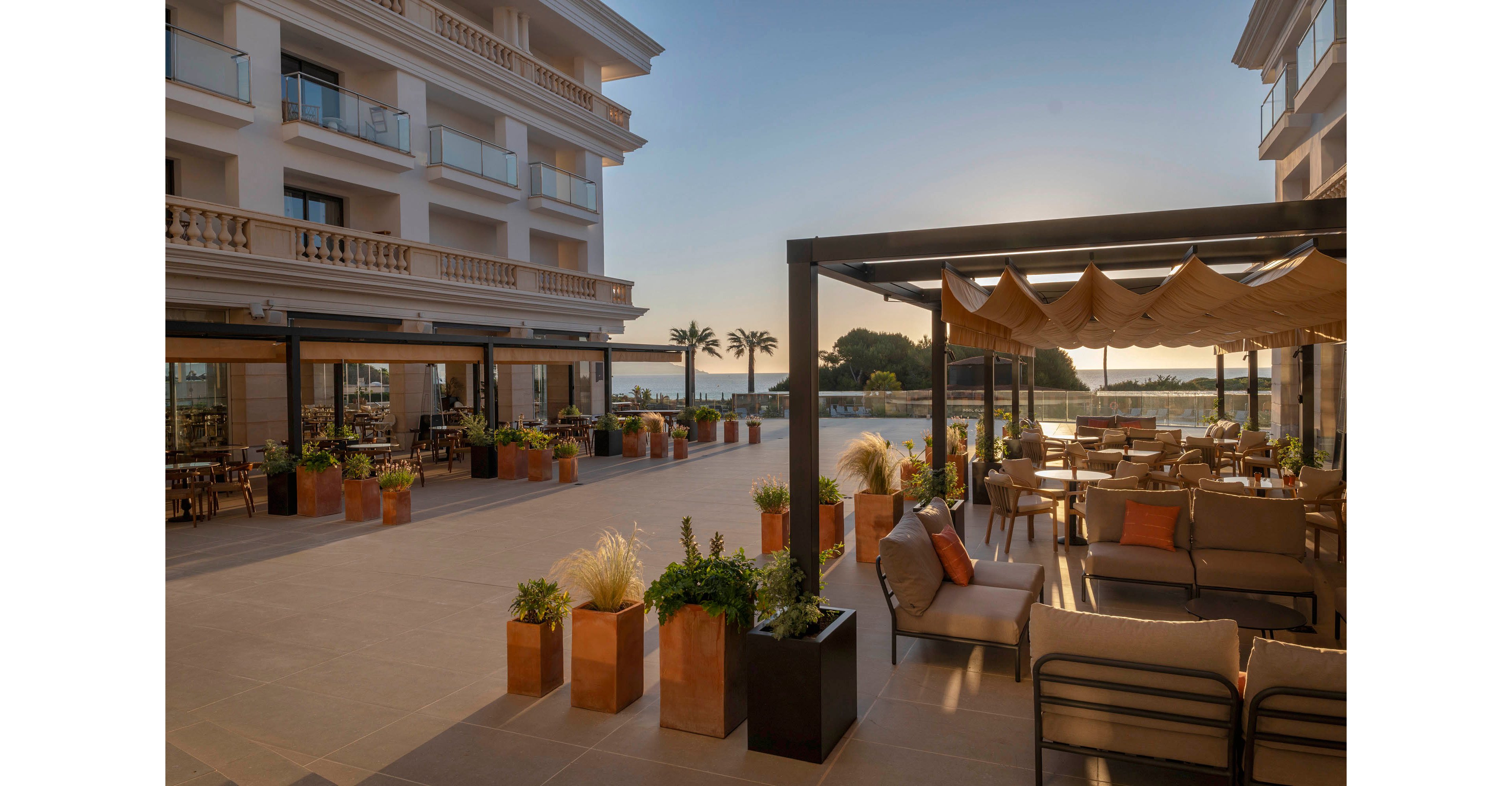An elegant hotel terrace lounge is bathed in warm evening sunlight. Comfortable outdoor seating with cushions is arranged beneath a modern pergola. Potted plants and greenery line the spacious tiled patio, adding a touch of nature. In the background, palm trees and calm water suggest a coastal or waterfront setting.