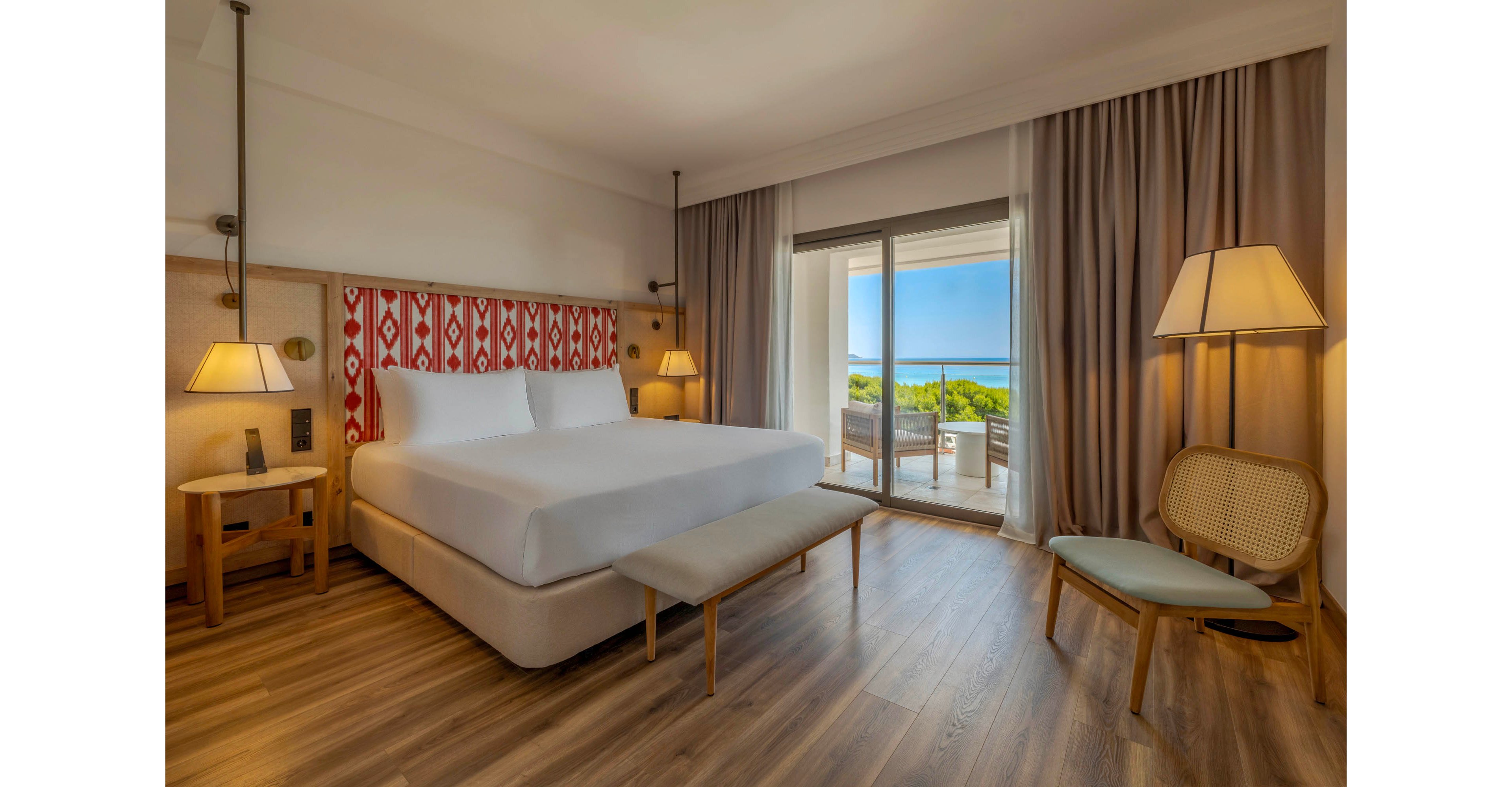 A spacious modern hotel bedroom features a large neatly made bed with white linens and a patterned red and white headboard. Warm wooden floors, matching bedside tables, and soft ambient lighting create a cozy atmosphere. A bench at the foot of the bed and a chair with a small table provide additional seating. Floor-to-ceiling curtains frame sliding glass doors that open to a balcony with outdoor furniture and a scenic ocean view in the distance.