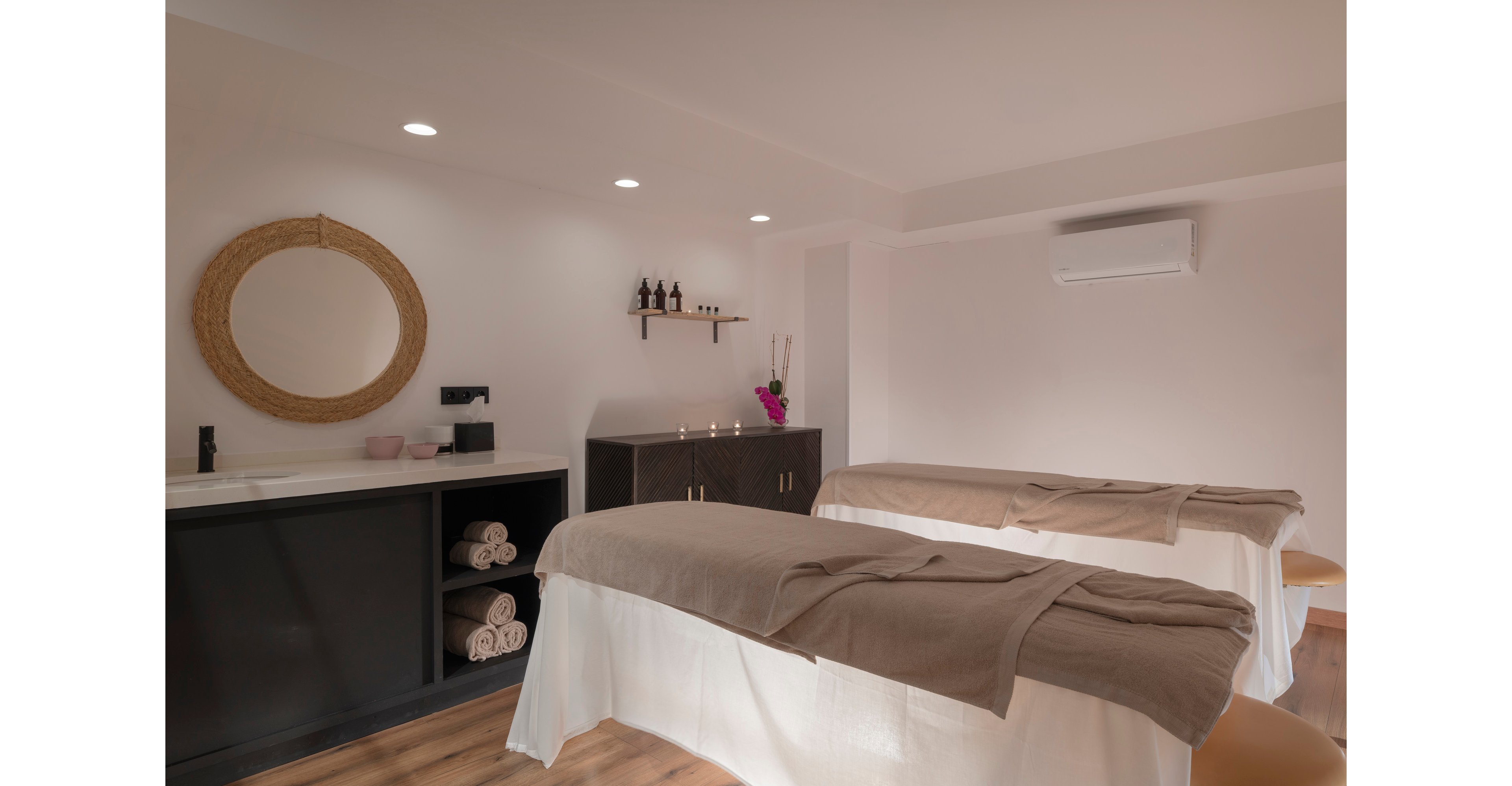 A serene spa treatment room features two massage tables covered with white linens and beige blankets. Soft, warm lighting creates a relaxing atmosphere, highlighting neatly rolled towels on open shelving. A round wall mirror, small bottles on a floating shelf, and candles add to the tranquil decor. The minimalist design and neutral color palette emphasize comfort and wellness.
