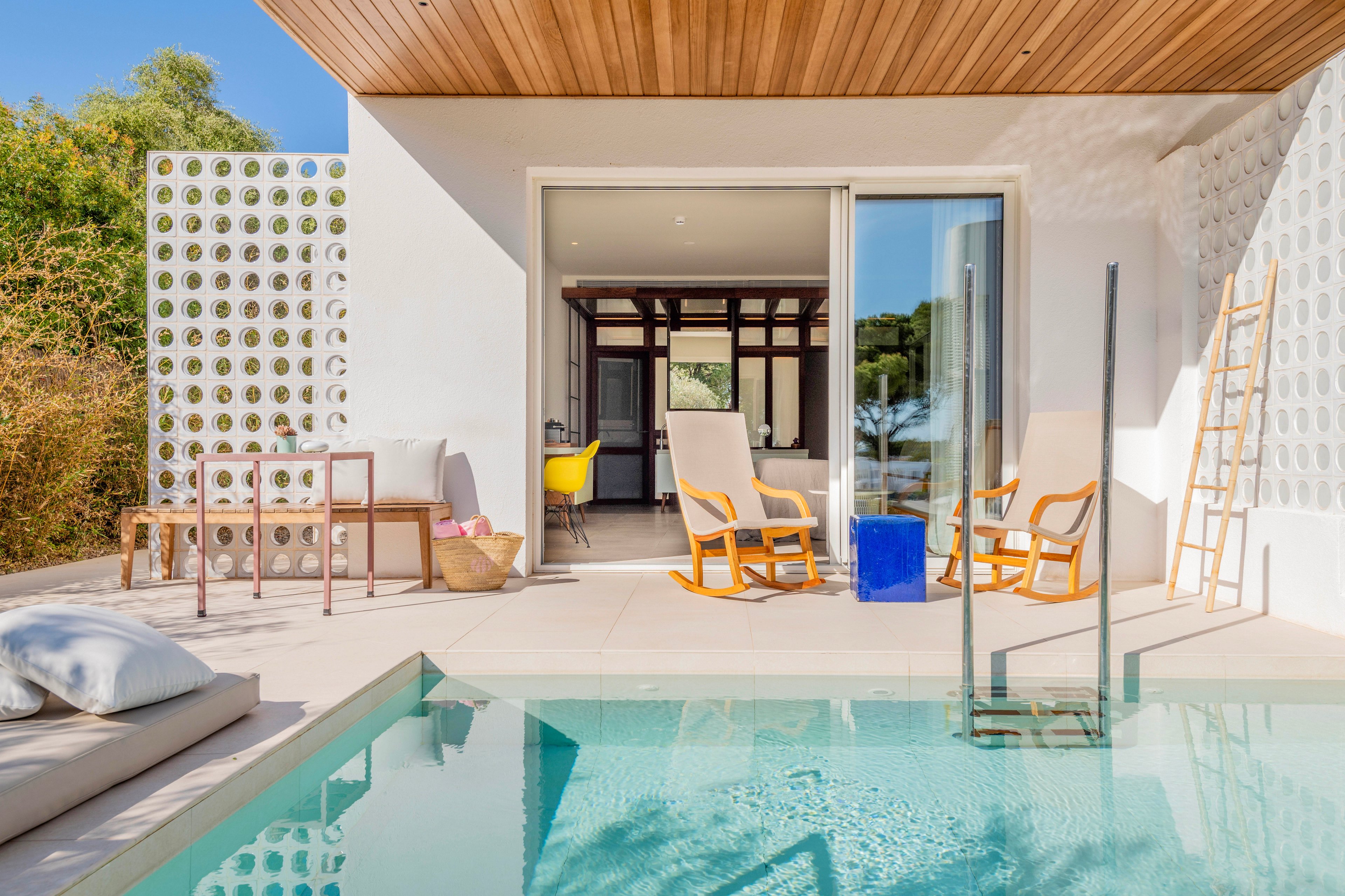 A contemporary outdoor patio opens directly onto a clear turquoise swimming pool. Wooden lounge chairs and a small table sit on a light stone terrace beside a glass pool barrier. The white house façade features circular cutout screens, a wooden ceiling, and large sliding glass doors leading to an interior with a yellow chair. Soft cushions, a woven basket, and a leaning wooden ladder add relaxed decorative touches. Bright daylight and clean lines create a calm, minimalist vacation atmosphere.