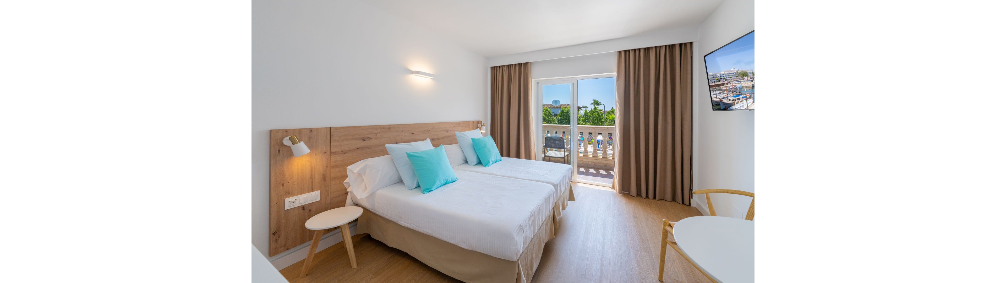 Modern hotel room featuring two twin beds with light blue accent pillows and wooden headboard. The space is bright and minimal, with neutral curtains framing a glass door that opens to a balcony. Outside, greenery and buildings are visible under a clear sky, adding a vacation feel. A wall-mounted television, small round tables, and simple chairs complete the clean, contemporary interior.