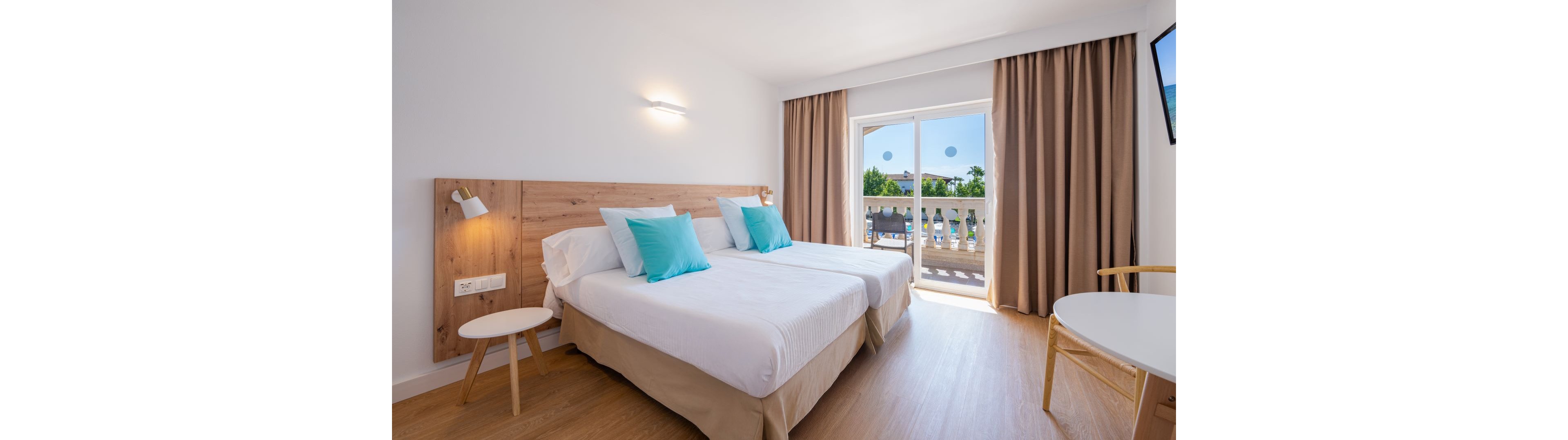 A bright, modern hotel room features two neatly made twin beds with white linens and light wood headboard. Turquoise accent pillows add a pop of color against the neutral beige bed skirts and curtains. Natural light floods in through the glass balcony doors, revealing an outdoor pool area and greenery. Minimalist furniture, including a small round table and chair, enhances the clean and relaxing atmosphere.
