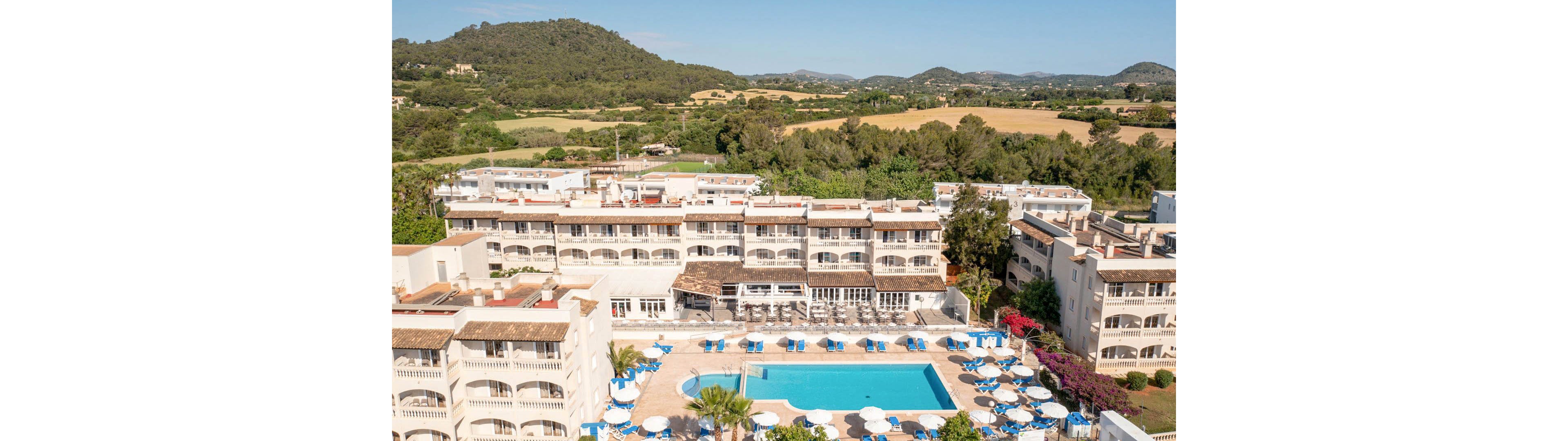 The image shows a large resort hotel complex with a central outdoor swimming pool surrounded by blue and white lounge chairs and umbrellas. The hotel buildings are white with terracotta roofs, set against a backdrop of green hills and open countryside. The setting is sunny and tranquil, suggesting a relaxing vacation atmosphere. No visible text or numbers are present in the image.