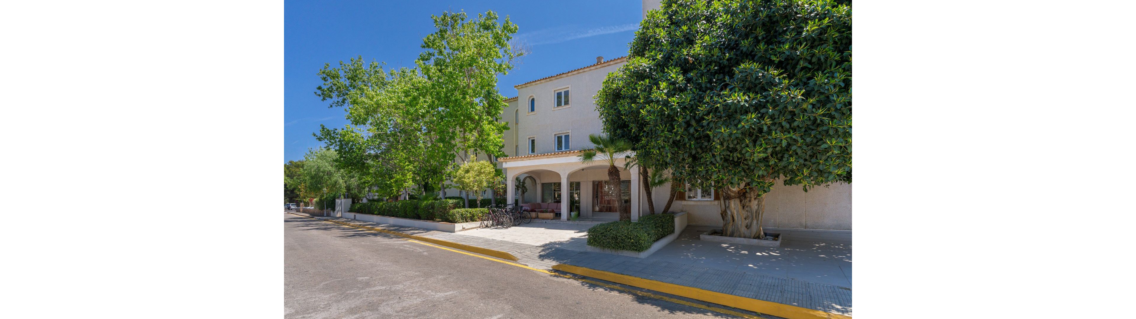 A bright, sunlit hotel building stands along a quiet street, surrounded by mature trees and manicured shrubs. The architecture features light-colored walls, arched entryways, and a tiled roof, evoking a Mediterranean style. Bicycles are parked near the entrance, and the scene is calm with clear blue skies overhead. No visible text or numbers are present in the image.