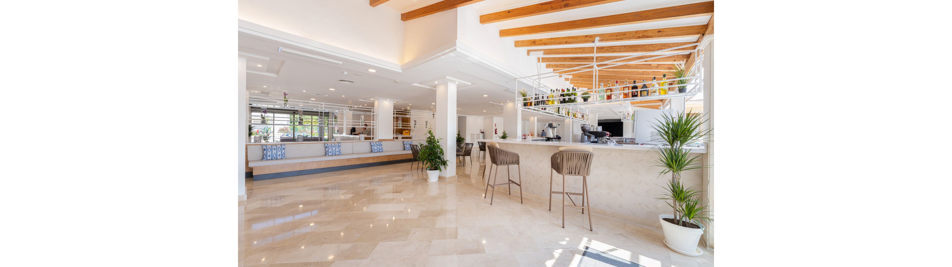 Spacious hotel lobby features a sleek bar area with high chairs and a variety of bottles displayed on shelves. The setting is bright and airy, with natural light streaming in and potted plants adding a touch of greenery. The interior design showcases a neutral palette with white walls, beige marble flooring, and wooden ceiling beams. No visible people are present, emphasizing the clean and inviting atmosphere. No explicit numbers or identifiable brands are visible in the image.