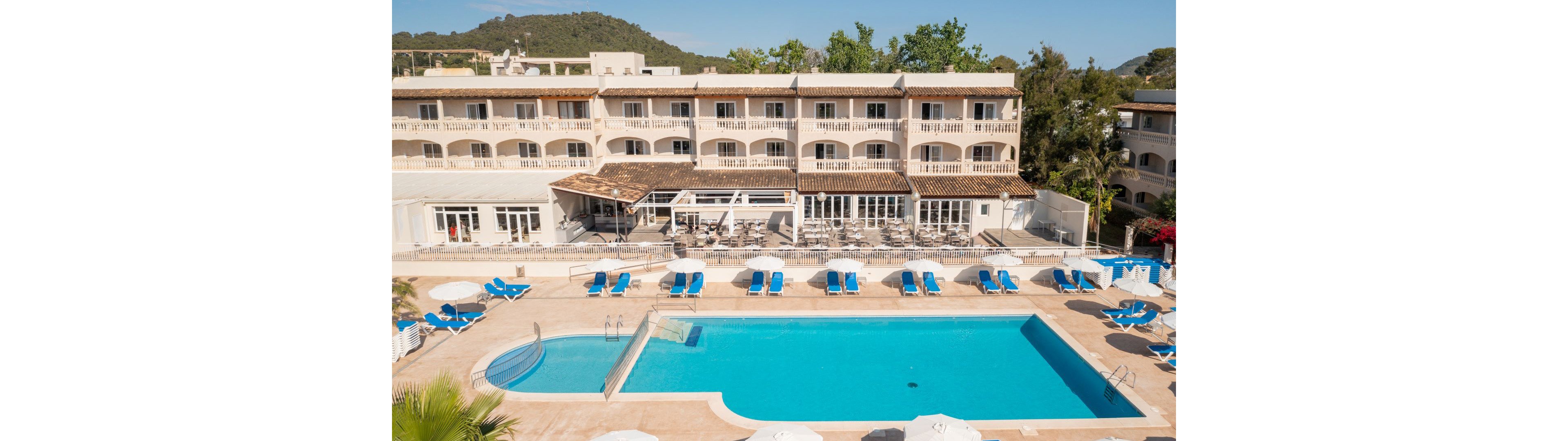 A large rectangular outdoor swimming pool is the central feature of this sunlit resort hotel. Blue lounge chairs and umbrellas are arranged around the pool, with a three-story Mediterranean-style building in the background. The setting is bright and inviting, with clear skies and lush greenery surrounding the area. No visible text or numbers are present in the image.